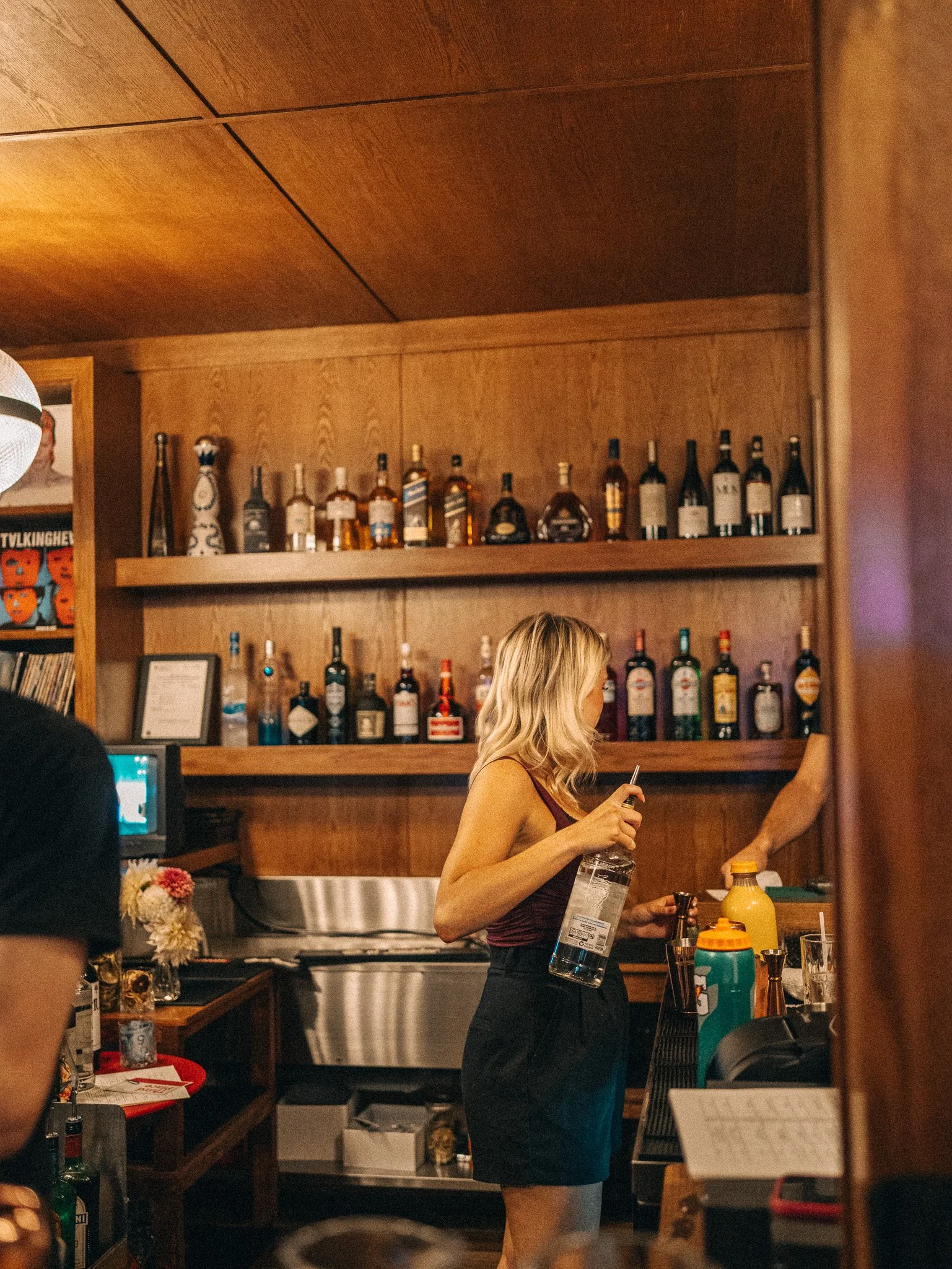 A woman with blonde hair wearing a sleeveless top prepares drinks behind a bar counter, with bottles of alcohol displayed on shelves behind her.