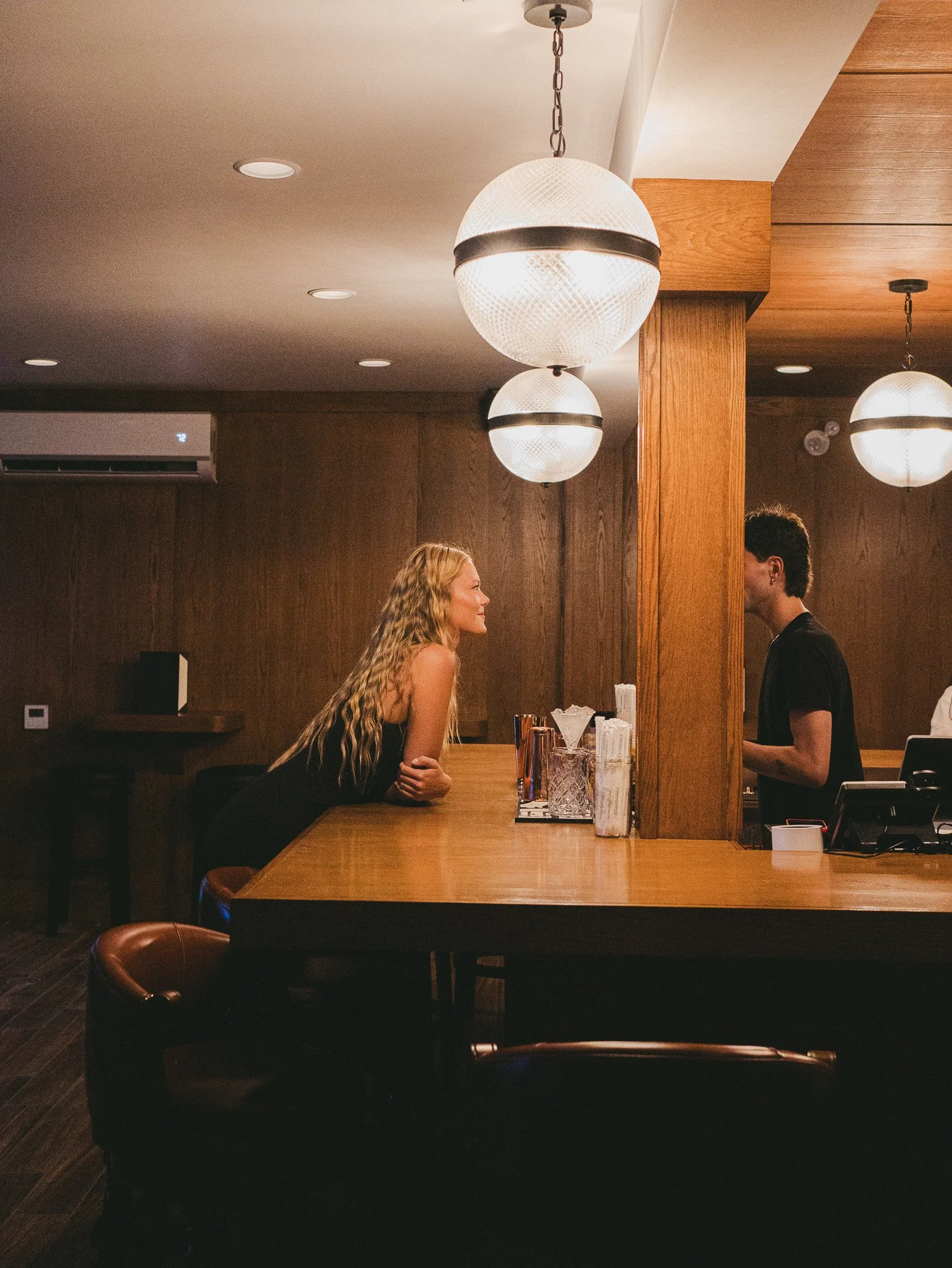 A woman with long blonde hair smiling and talking to a man at a bar or counter in a dimly lit restaurant or cafe with wooden decor and large hanging light fixtures.