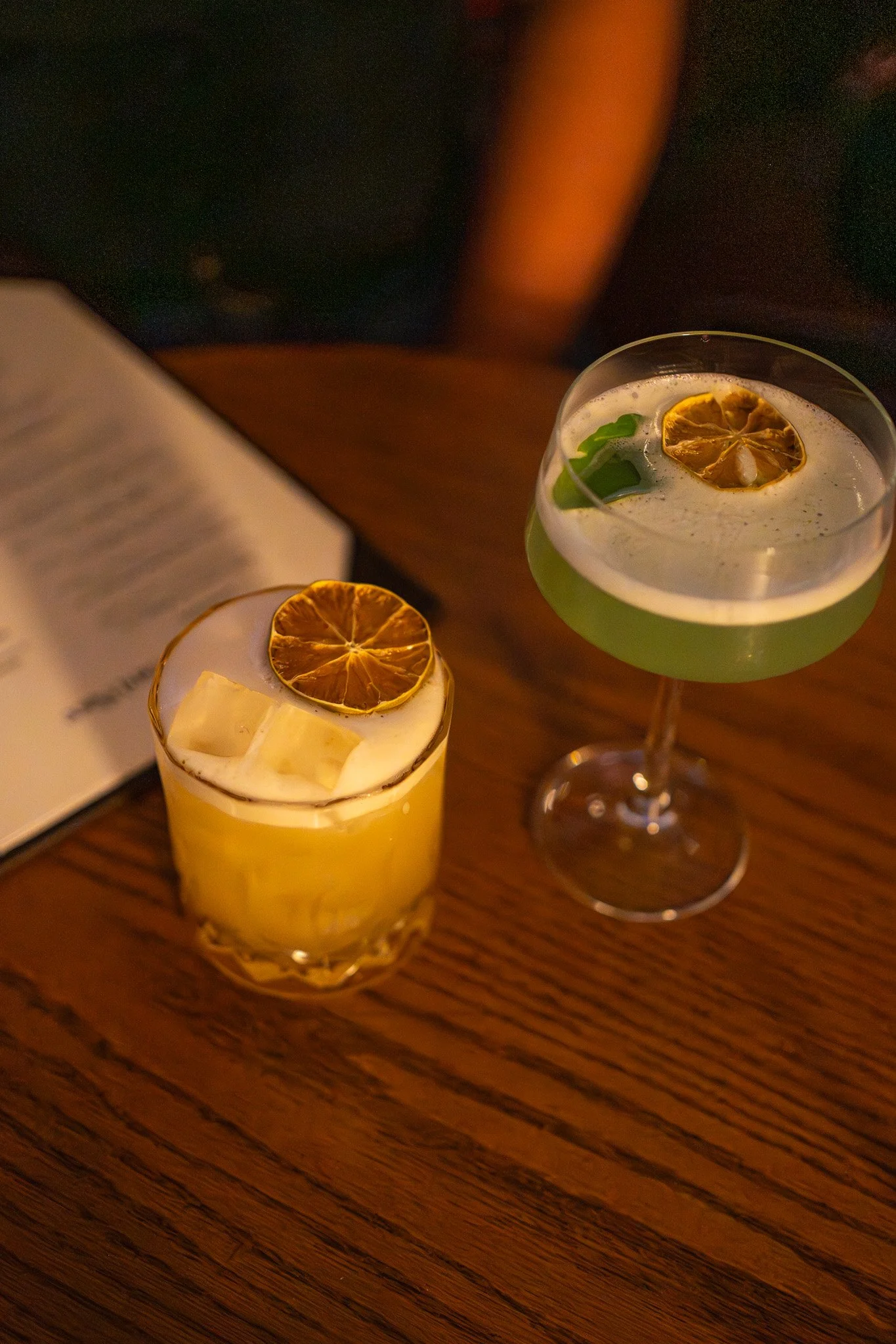 Two cocktails with dried lemon slices on top, one in a short glass with ice, the other in a tall glass with green garnish, on a wooden table.