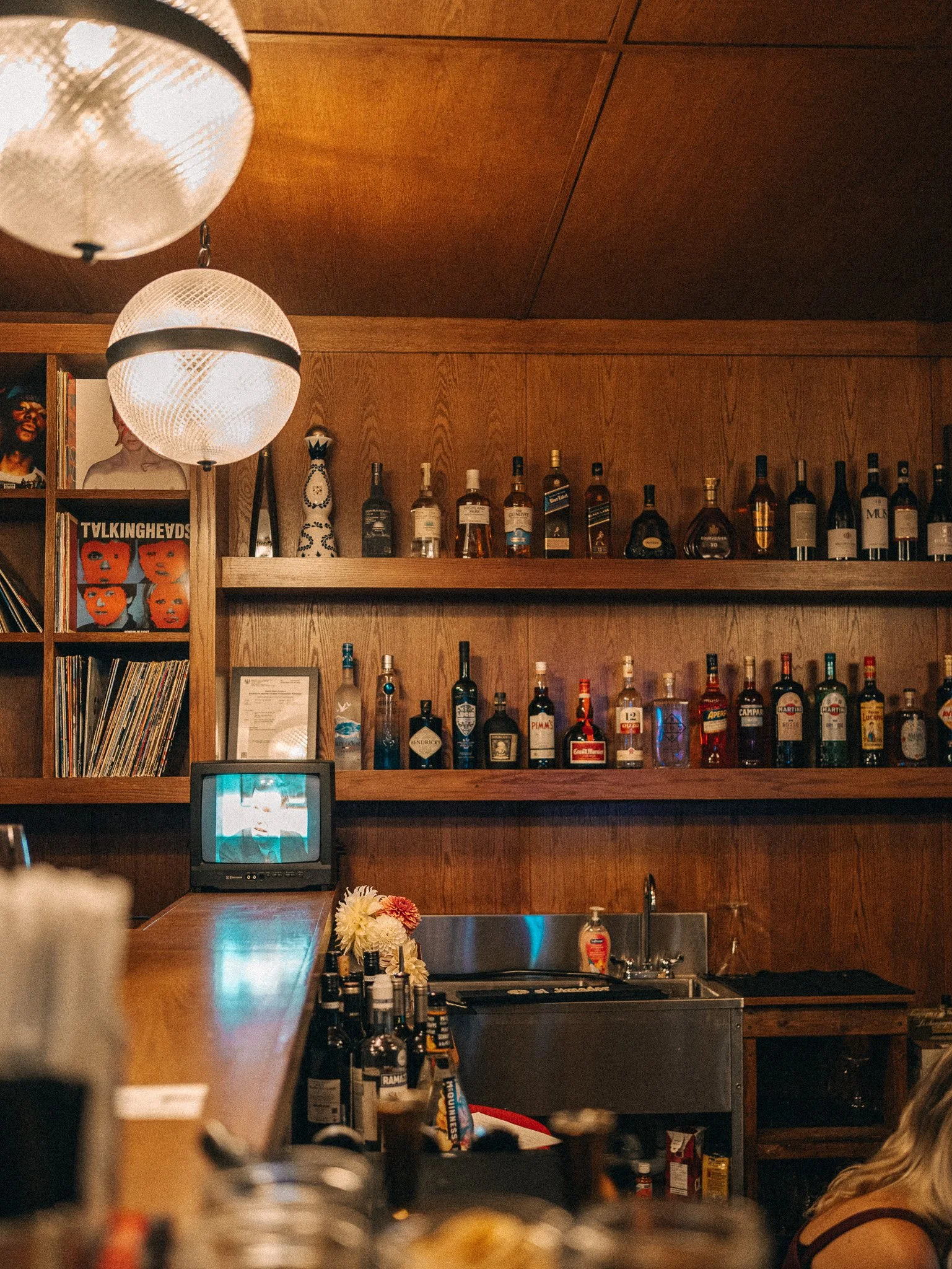 Bar area with wooden shelves holding bottles of liquor, a small TV showing a scene, a sink, a bouquet of flowers, and two hanging white lamps with black bands attached to a wooden ceiling.