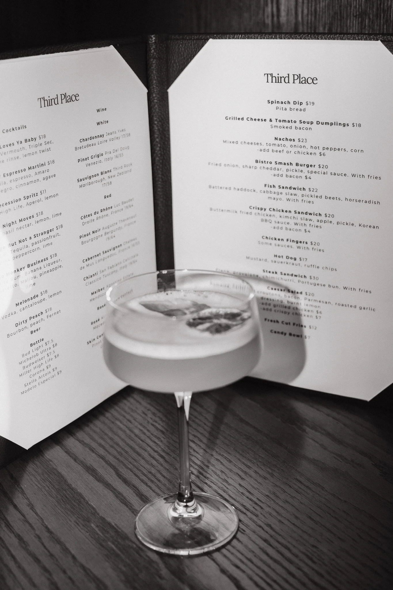 A glass of white wine on a dark wood table with menus in the background listing various food and drink options and prices.