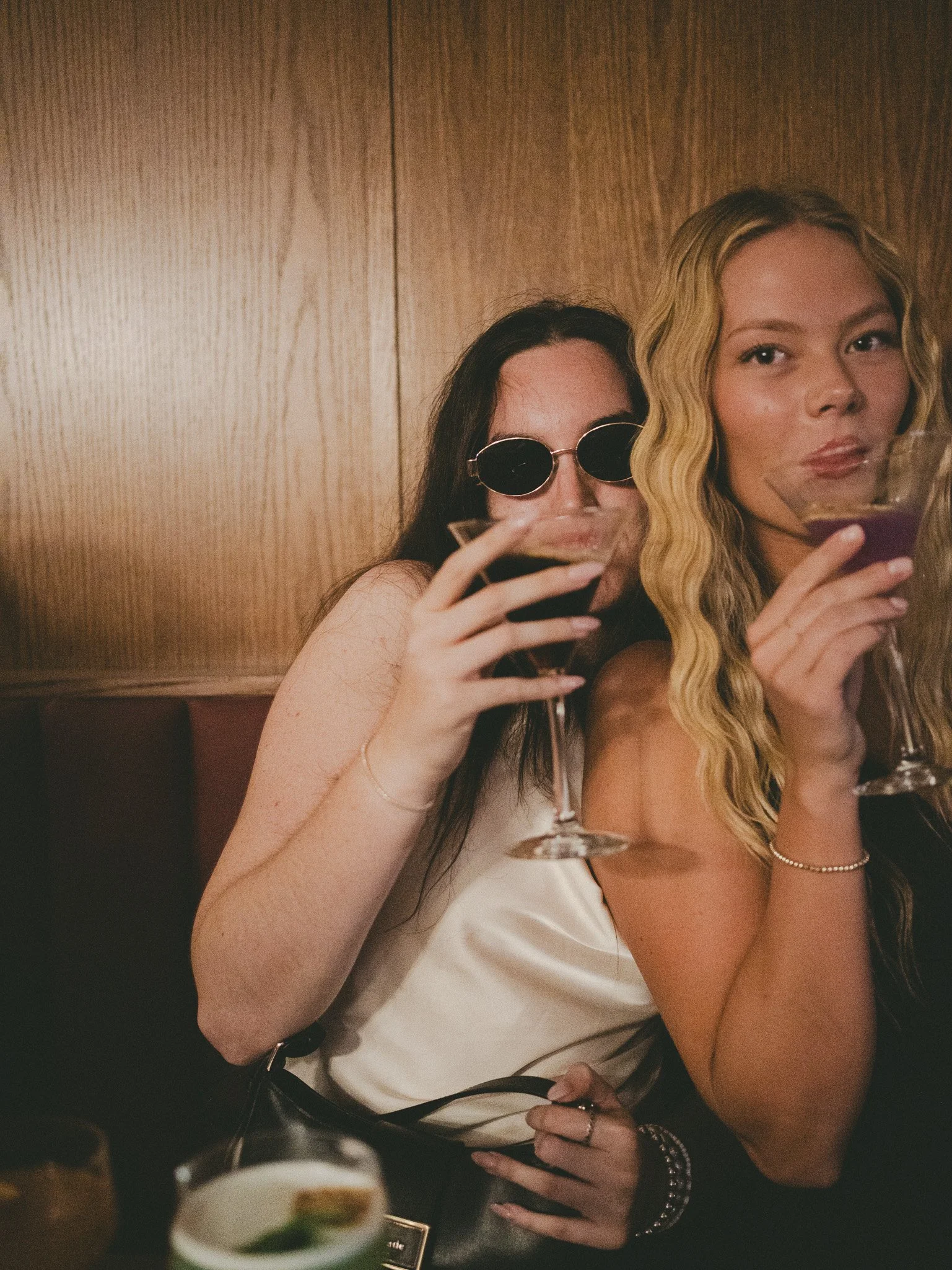Two women are sitting together enjoying drinks in a bar or restaurant. One woman has long, wavy blonde hair and is wearing minimal makeup, a bridal gown, and jewelry. The other woman has long, dark hair, and wears dark sunglasses. Both women are hold