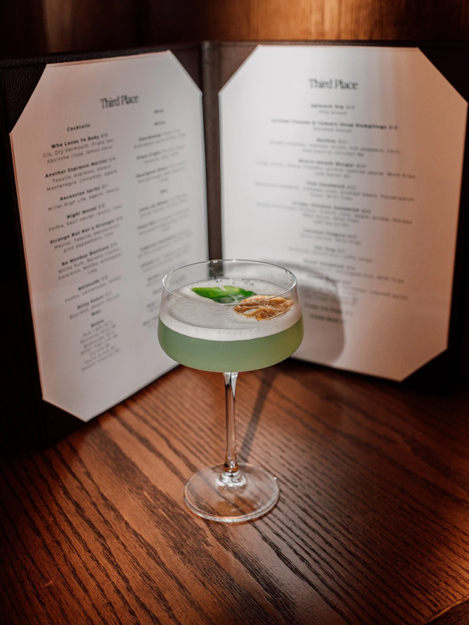 A green cocktail in a coupe glass garnished with a mint leaf and a sliced fruit, placed on a wooden surface with an open menu in the background.