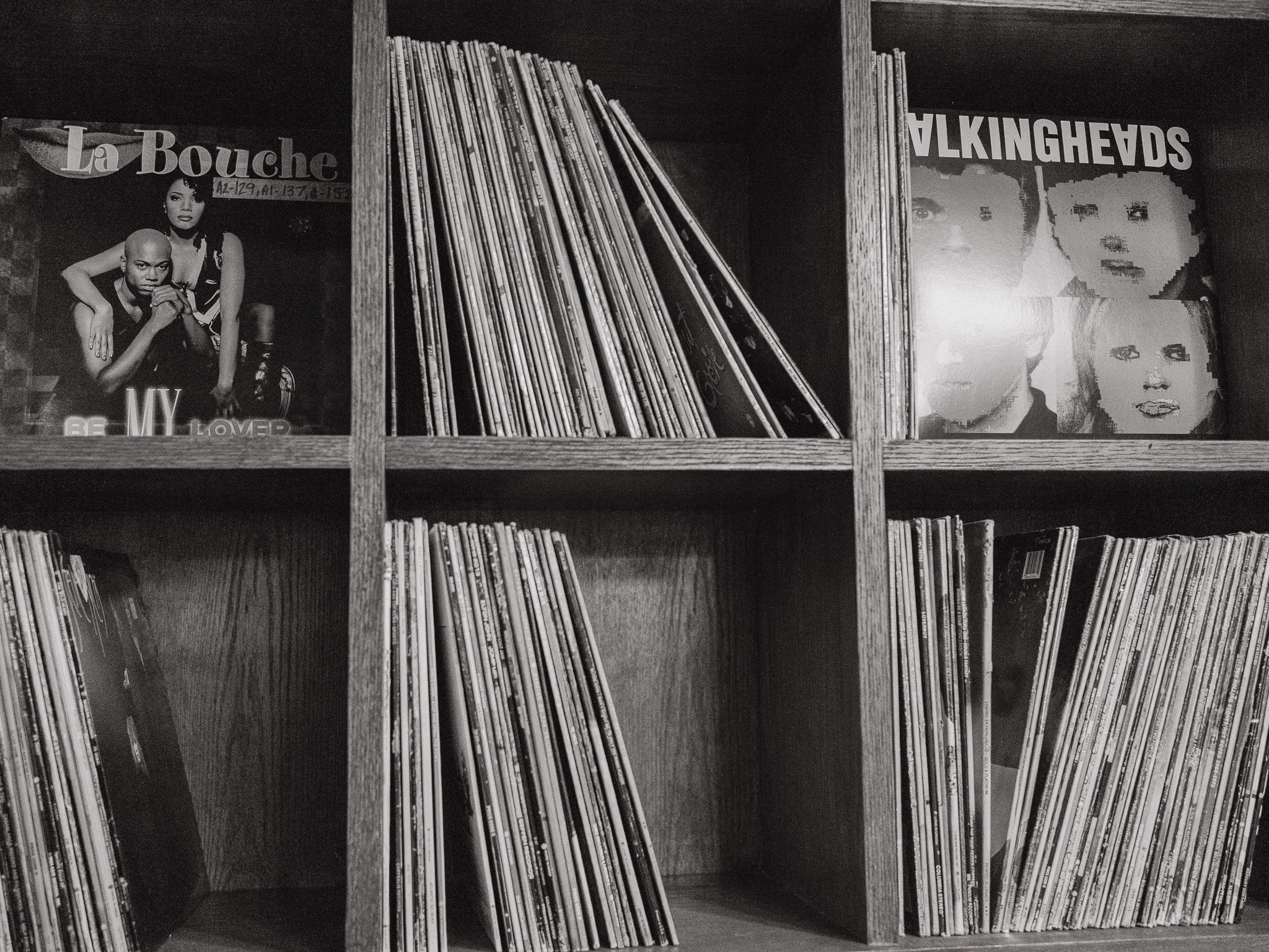 Black and white photo of a wooden bookcase filled with vinyl records on shelves. Some records are leaning, and two album covers are visible on the top shelf, featuring graphic and photographic designs.