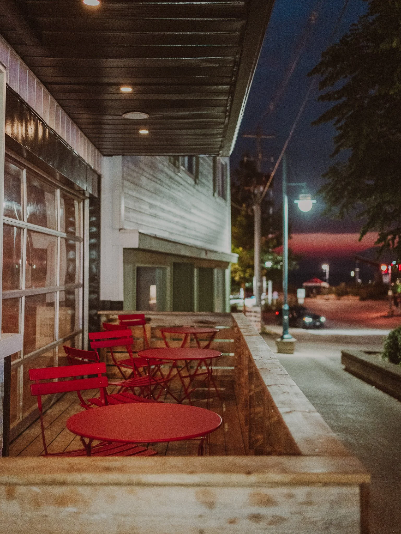 Nighttime view of a small outdoor seating area with red tables and chairs on a wooden deck outside a building, illuminated by overhead lights. Street and utility poles, trees, and a darkening sky with a hint of sunset colors are visible in the backgr