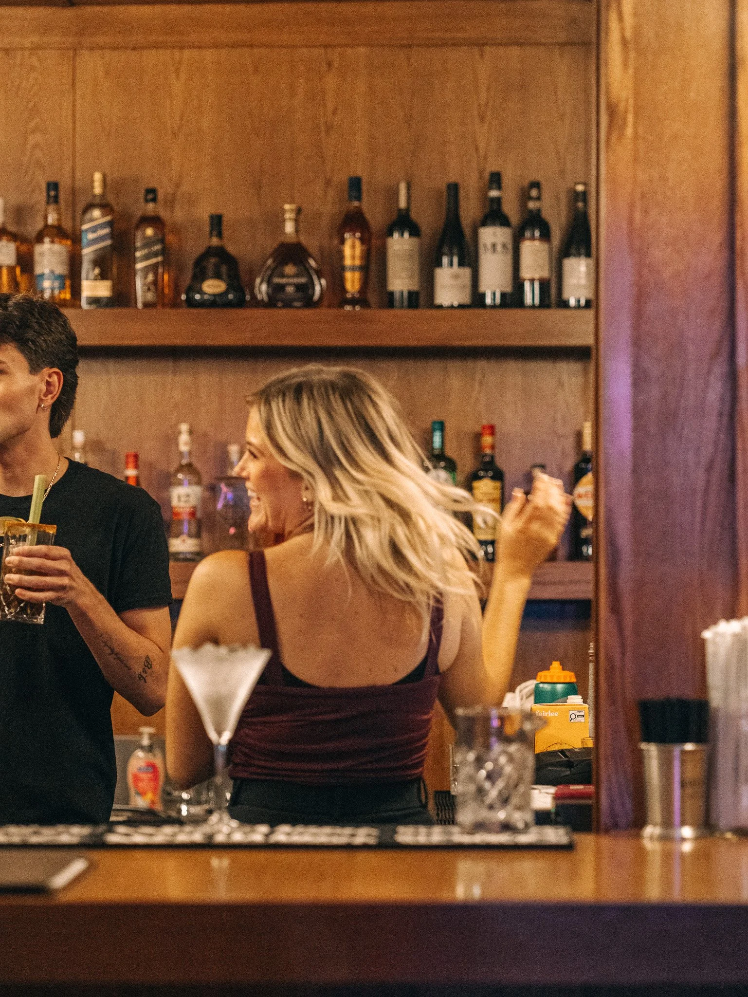 Reflection of a woman with blonde hair and a man with dark hair at a bar or restaurant, with shelves of liquor bottles in the background.
