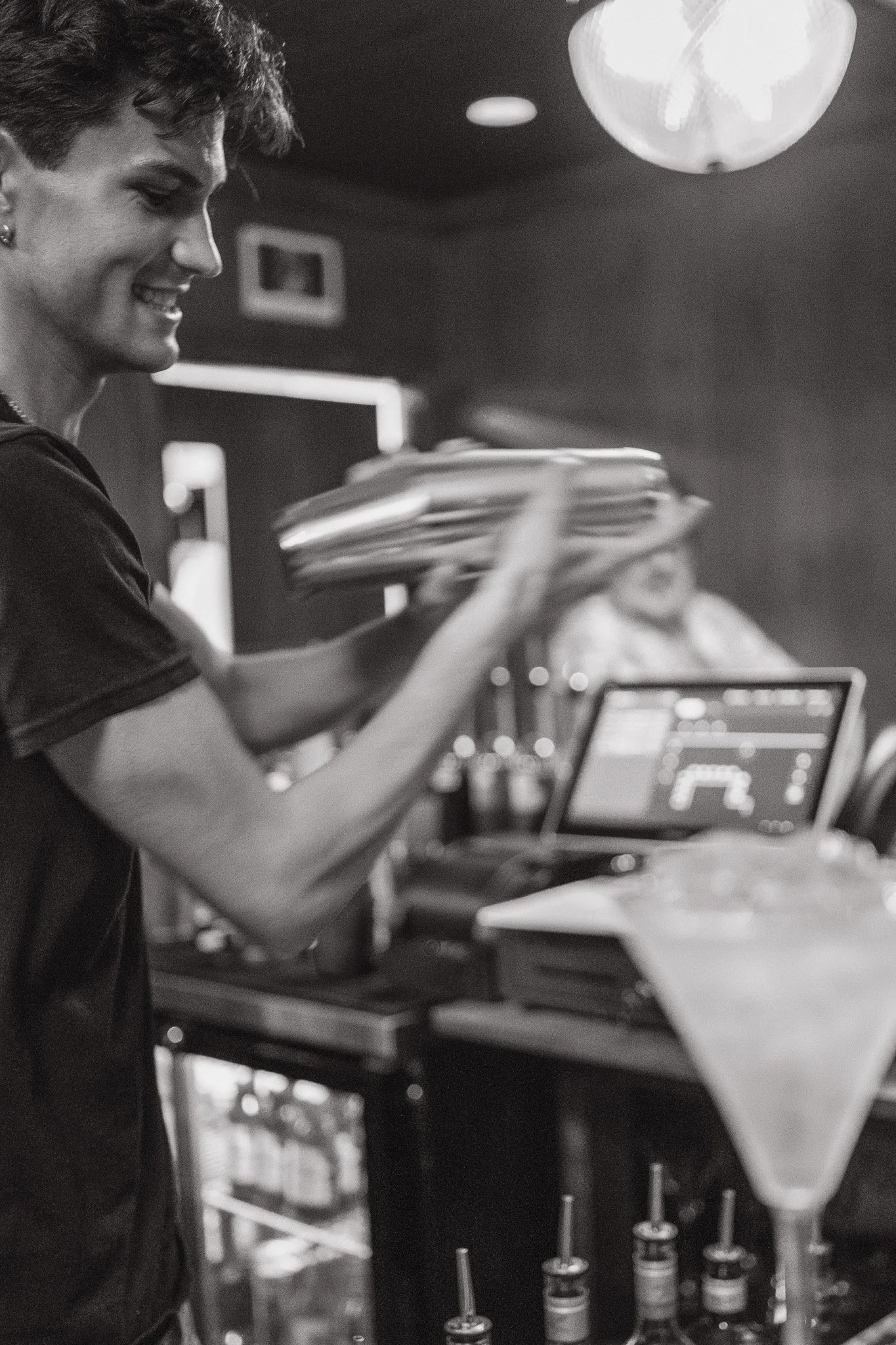 A woman smiling, working on a laptop at a bar or café, with a paper bag and some bottles on the table and a large hanging light fixture overhead.