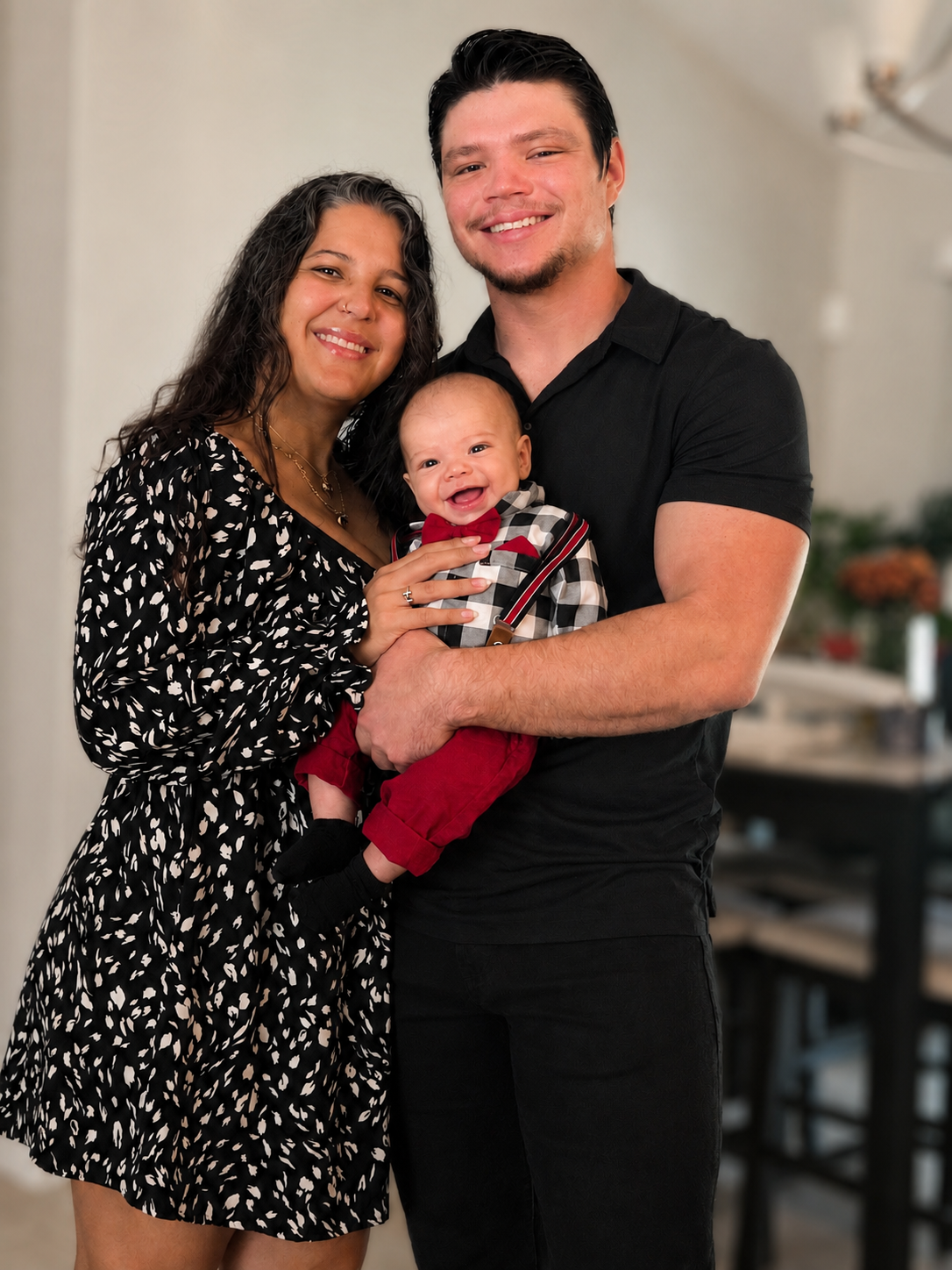 A smiling family of three, including a woman, a man, and a baby, standing indoors; the woman has curly dark hair, the man has short dark hair, and the baby is wearing a red, black, and white checkered shirt with a red bow tie.