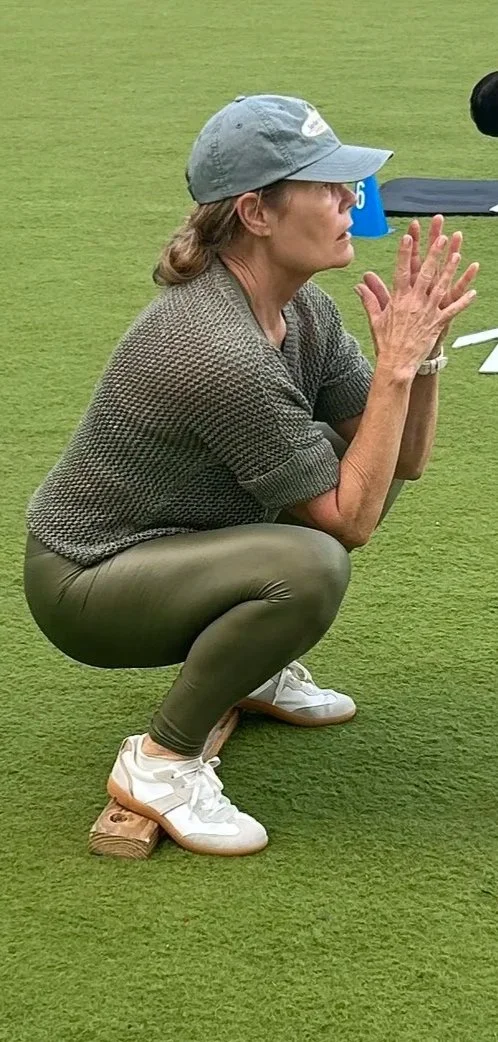 A woman squatting outdoors on grass, wearing a gray cap, brown leggings, a brown sweater, and white sneakers, with an unusual shoe-on-wooden-block setup.