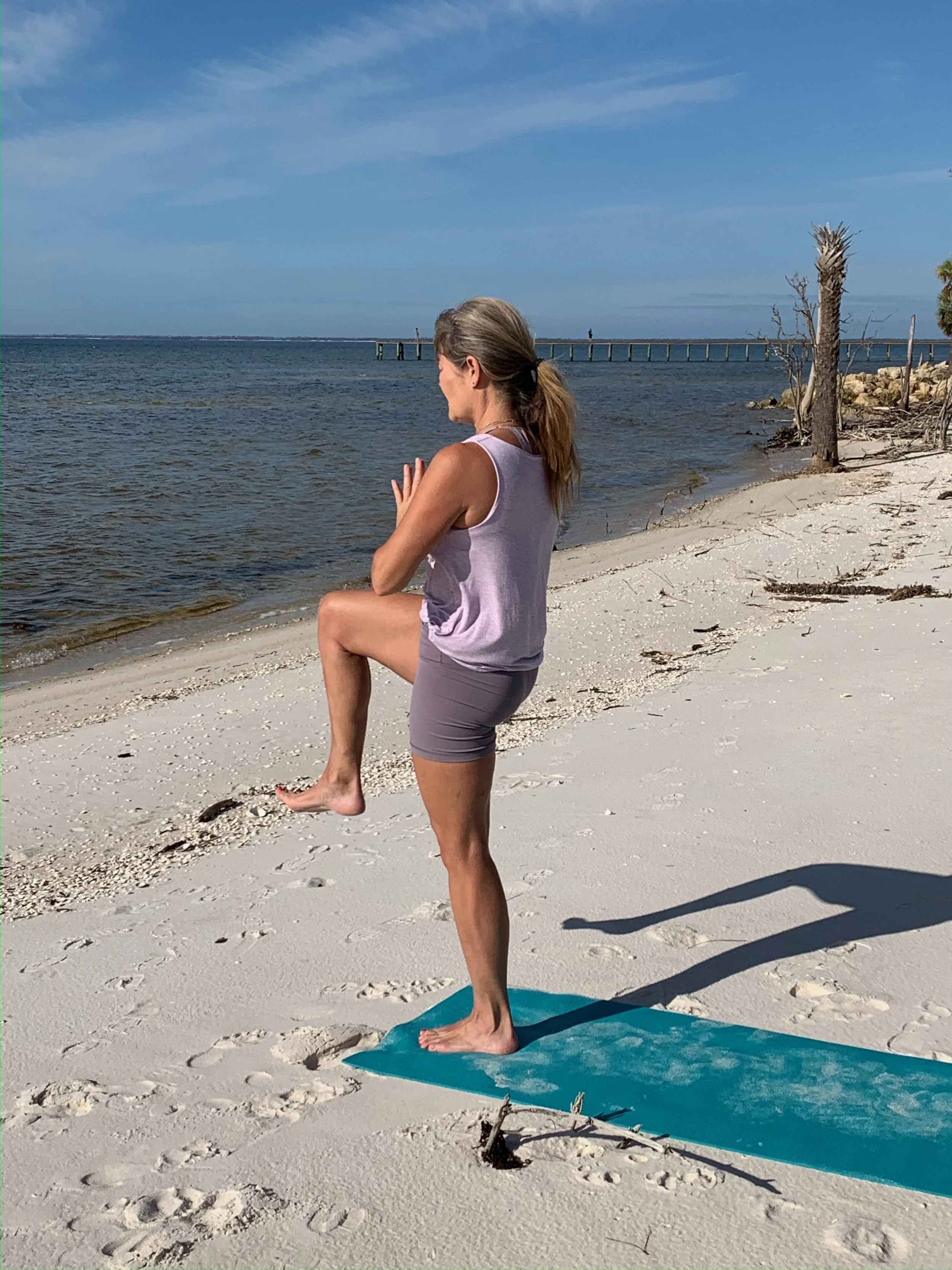 A woman practicing yoga on a blue mat on a sandy beach, performing a tree pose overlooking the ocean with a pier in the distance, under a clear blue sky.