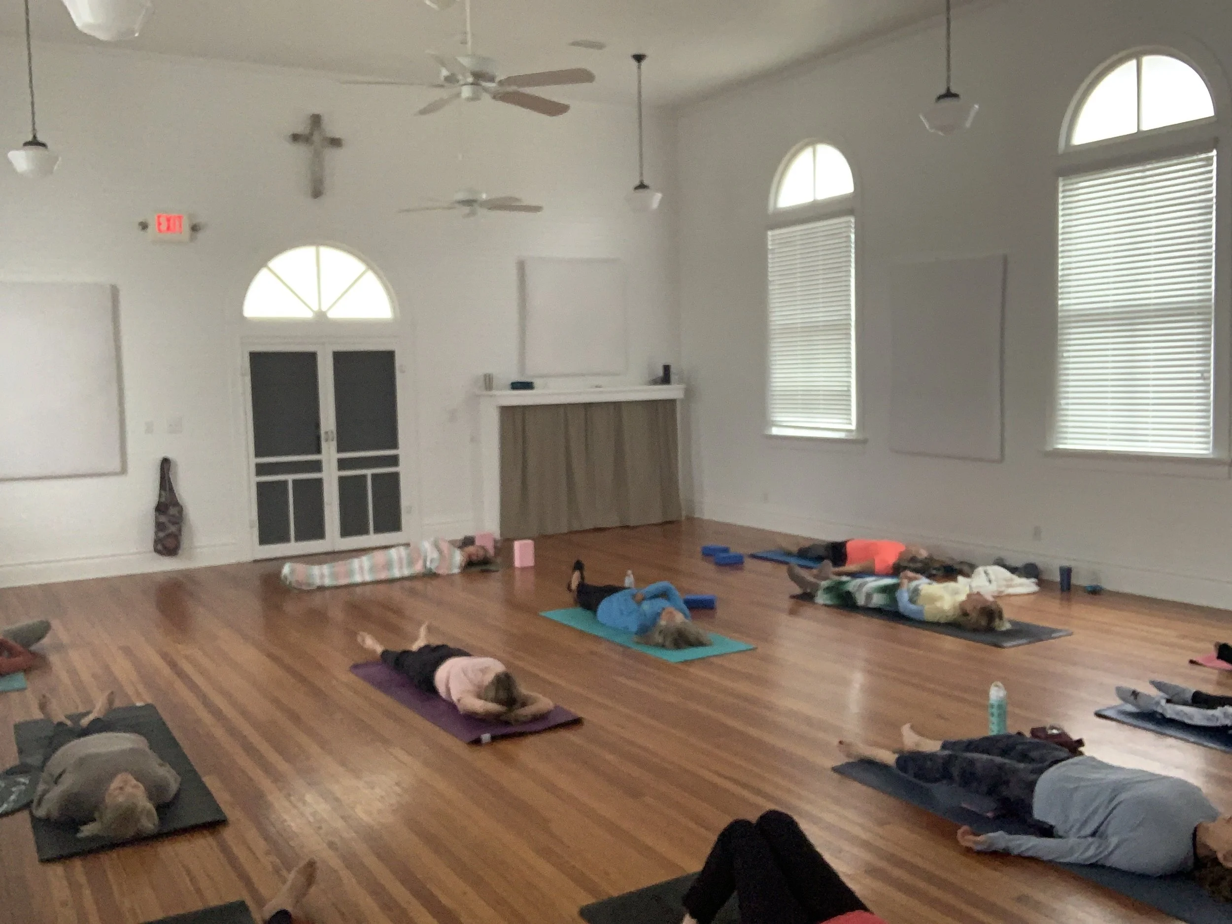 People lying on yoga mats in a church hall with large arched windows, ceiling fans, and a cross on the wall, participating in a group yoga or relaxation class.