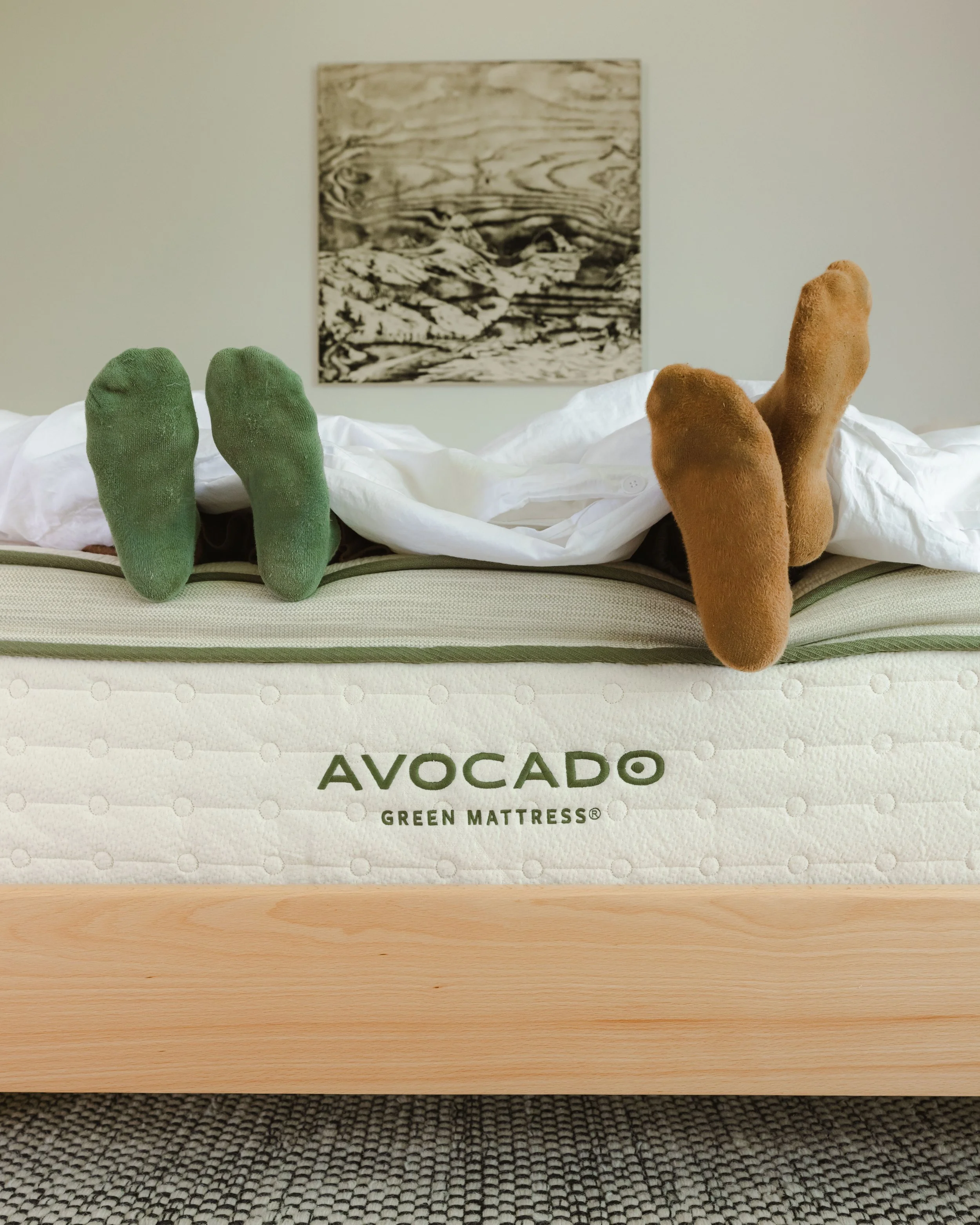 Two pairs of feet, one with green socks and one with brown socks, sticking out from a bed on top of a mattress labeled "AVOCADO Green Mattress." The mattress is on a wooden bed frame, and behind it is a beige wall with a landscape black-and-white pic