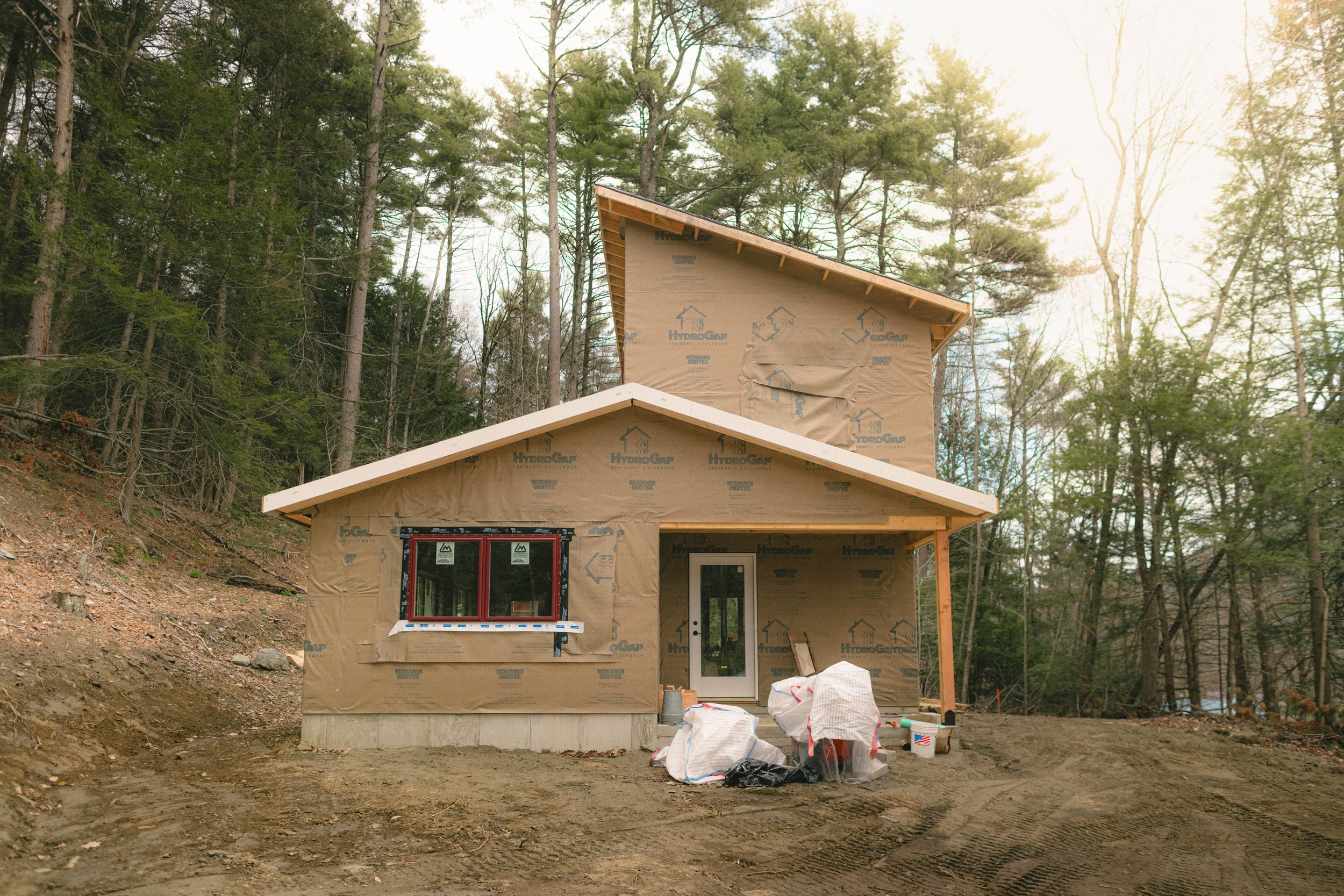 A house under construction with beige house wrap paper, surrounded by trees, on a dirt plot with construction materials and bags of supplies in front.