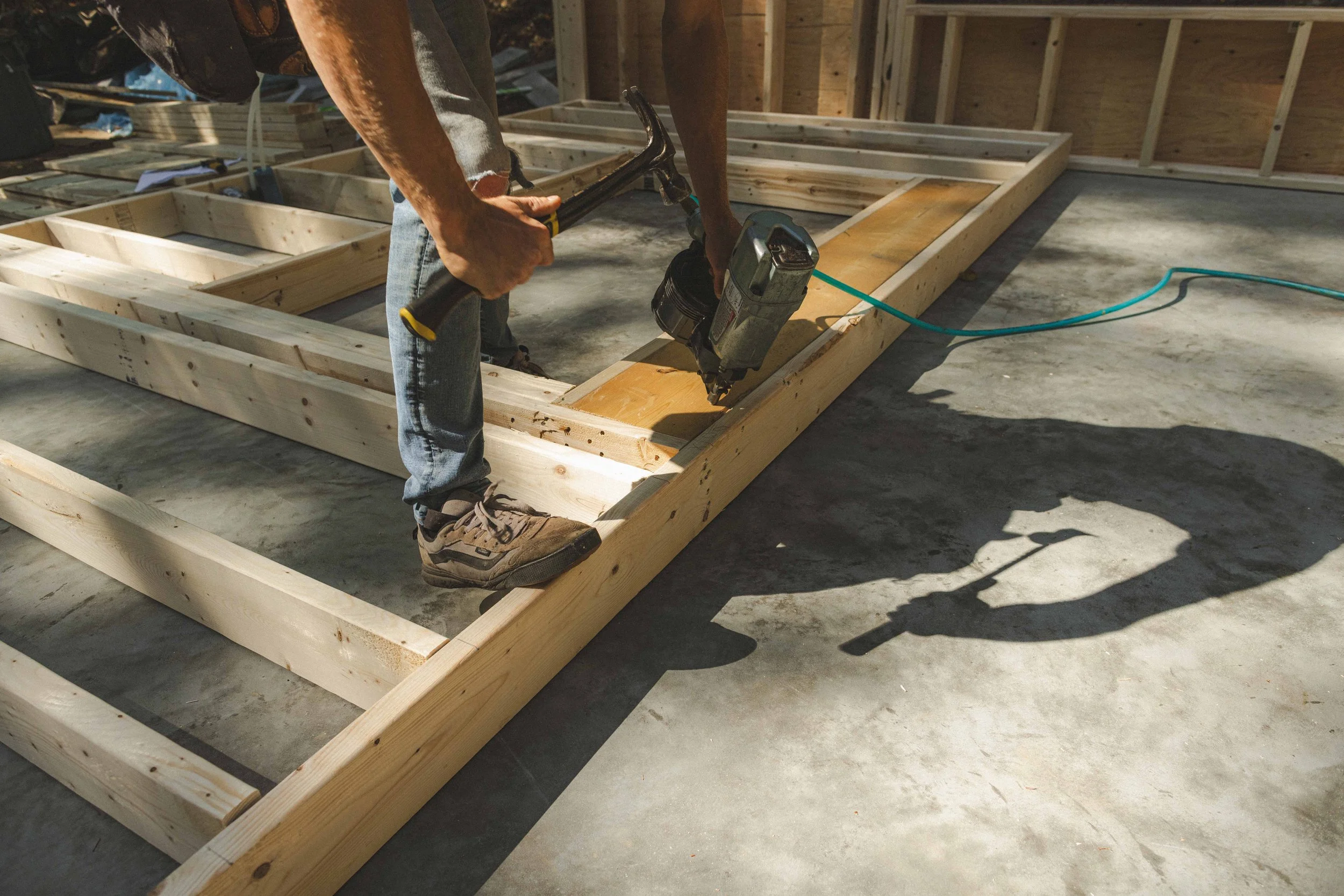 A person using a hammer and a nail gun to assemble wooden frames on a concrete floor inside a construction site.