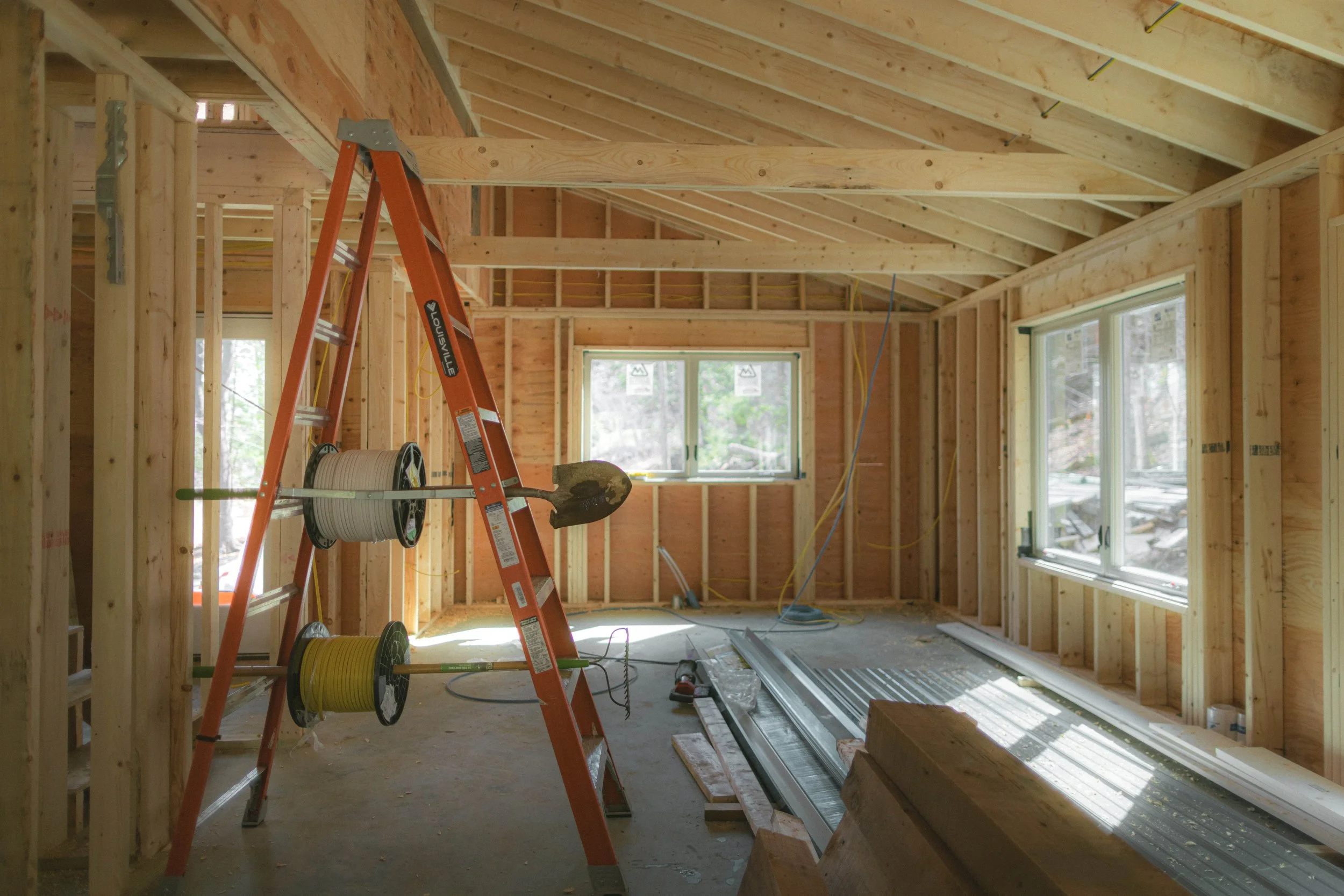 Interior view of a house under construction with exposed wooden framing, ladder, power tools, and construction materials.