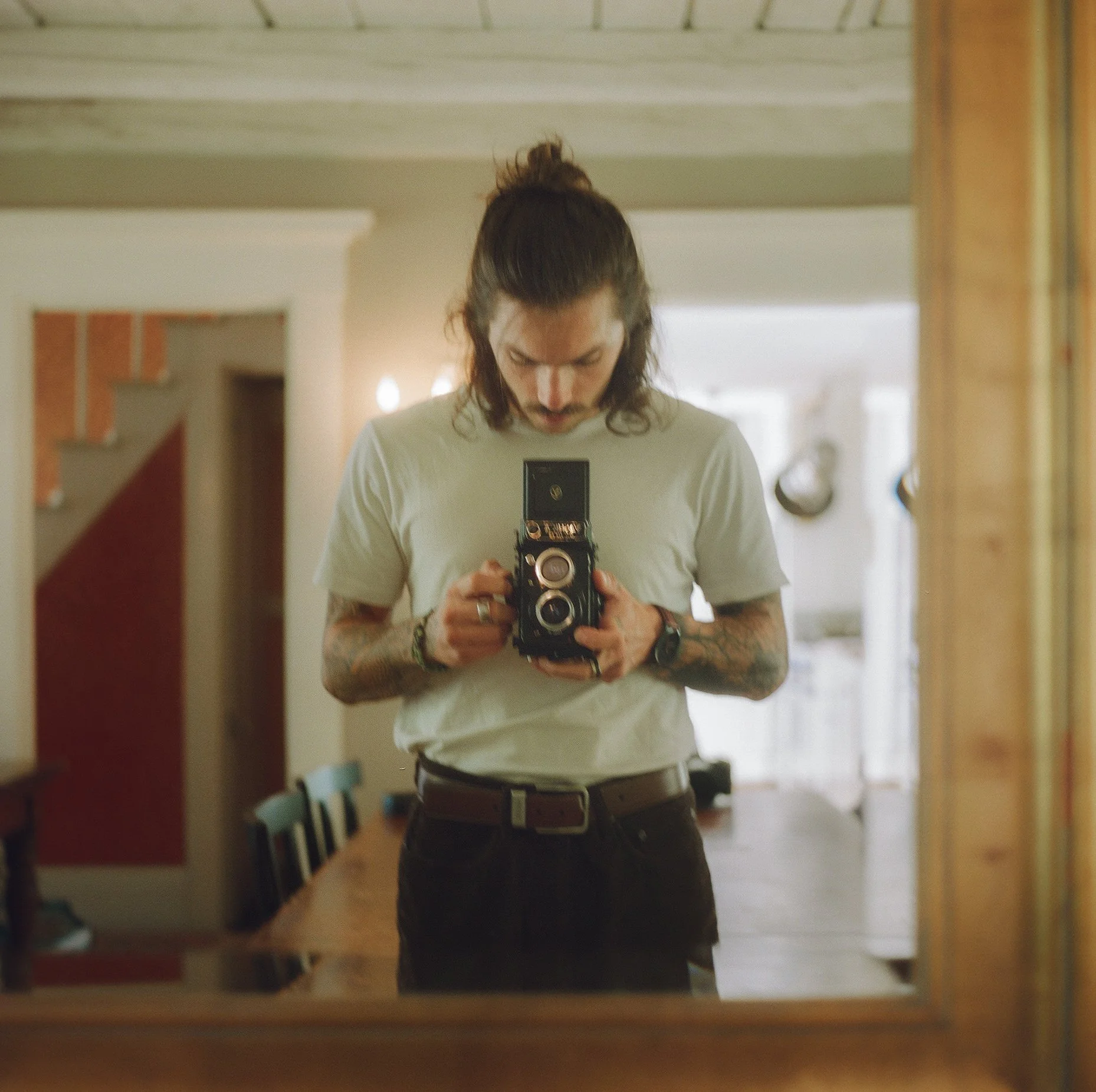 A man with long hair and tattoos taking a selfie in a mirror while holding a camera, standing in a room with chairs and a table.