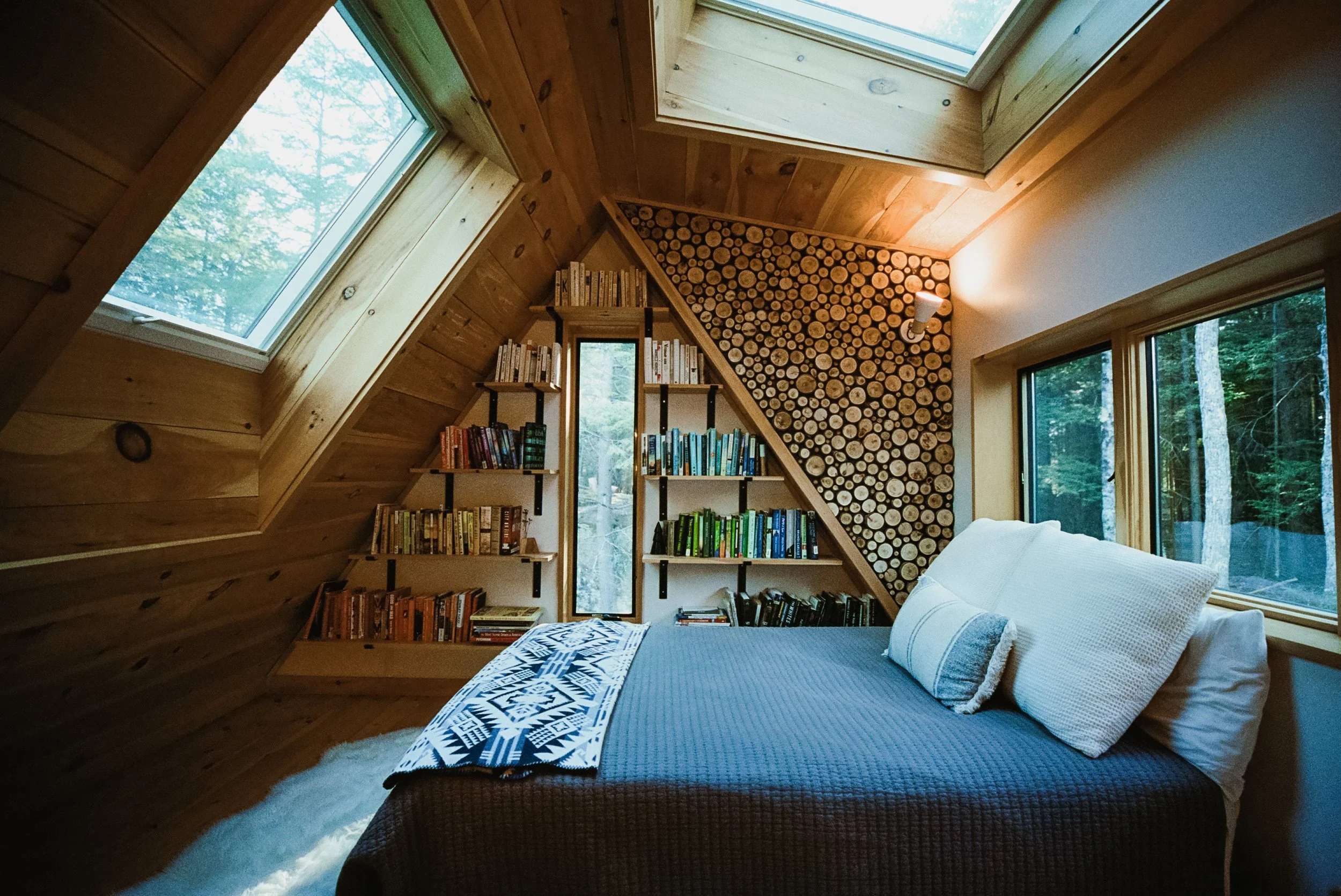 A cozy bedroom with wood-paneled sloped ceiling and walls, large windows showing a forest view, a bed with a white blanket and throw pillows, bookshelves filled with books, a wall with stacked logs, and a wall-mounted lamp.