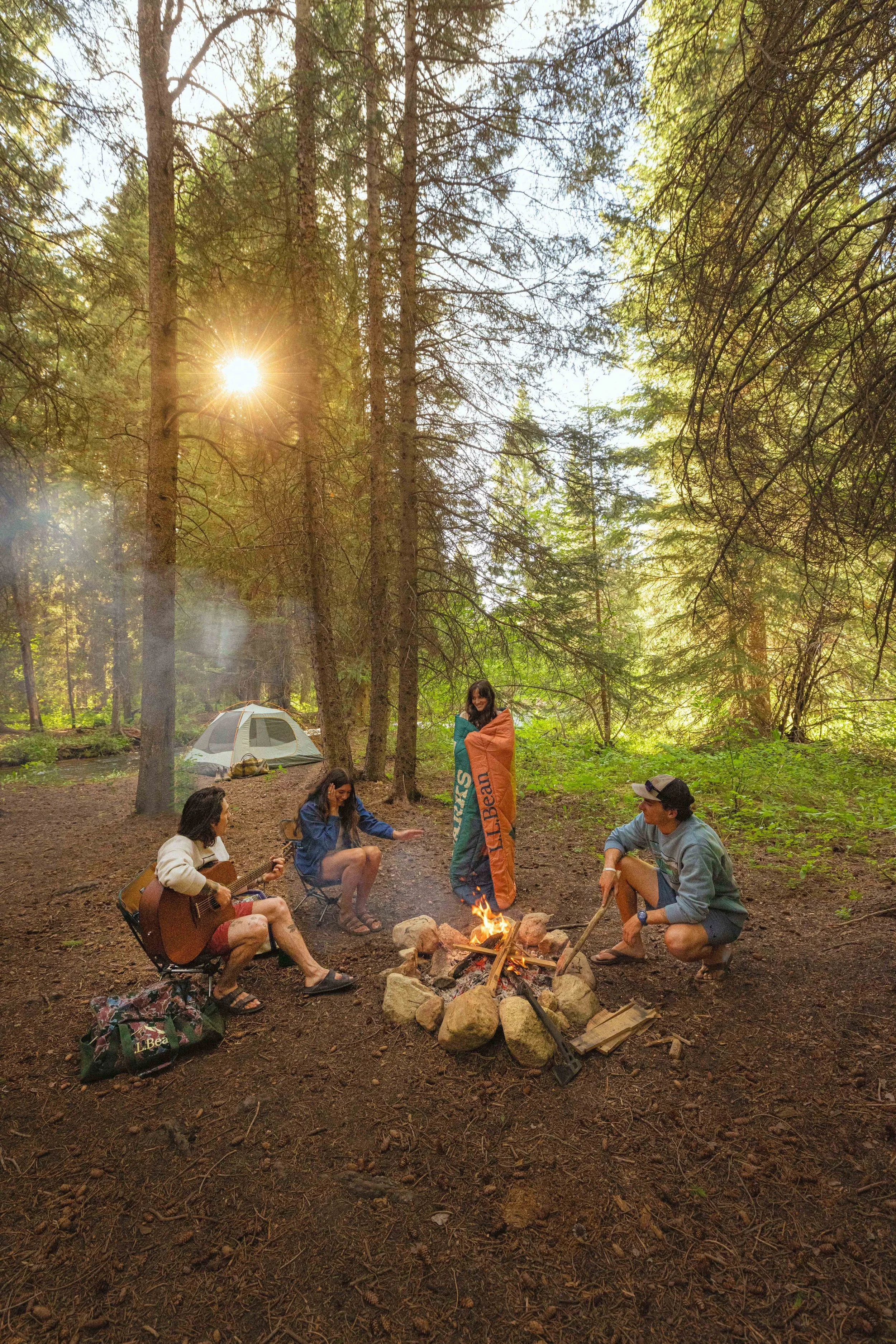 A group of people gathered around a campfire in a forest, some playing guitar and others sitting and chatting, with tents set up in the background, sunlight filtering through the trees.