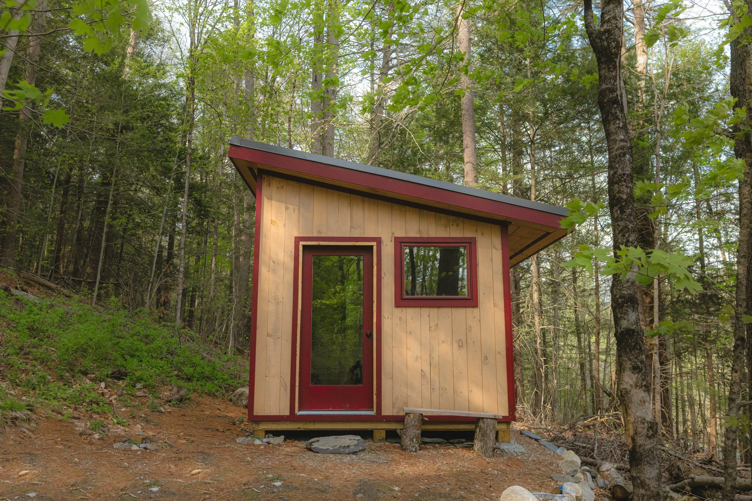 Small wooden house with red trim and a sloped gray roof, situated in a forest with tall trees and green foliage.