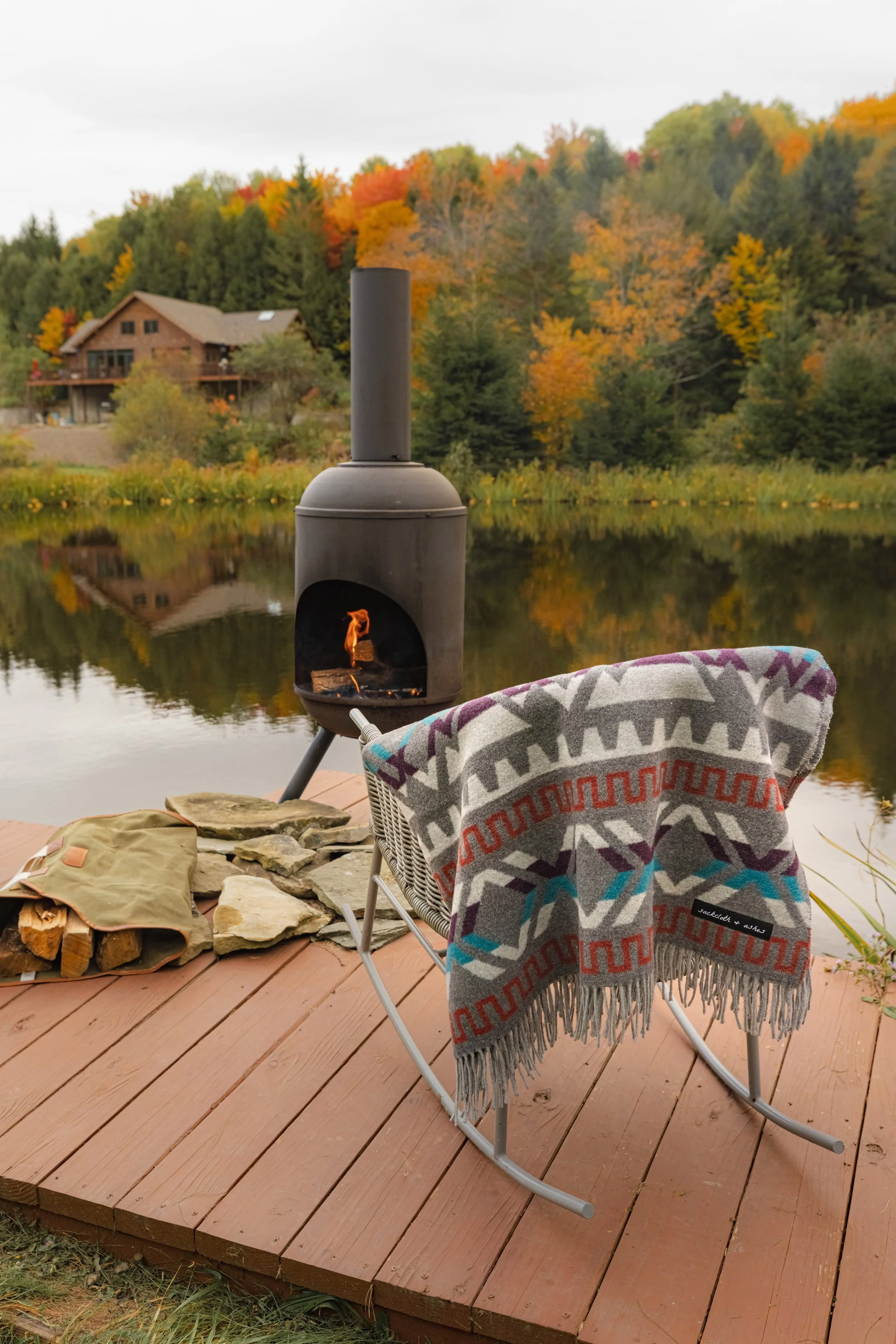 A cozy outdoor scene by a lake during autumn with a small fire pit on a wooden deck, a patterned blanket on a metal chair, a stack of firewood, a blanket, and a background of fall trees and a house reflecting in the water.