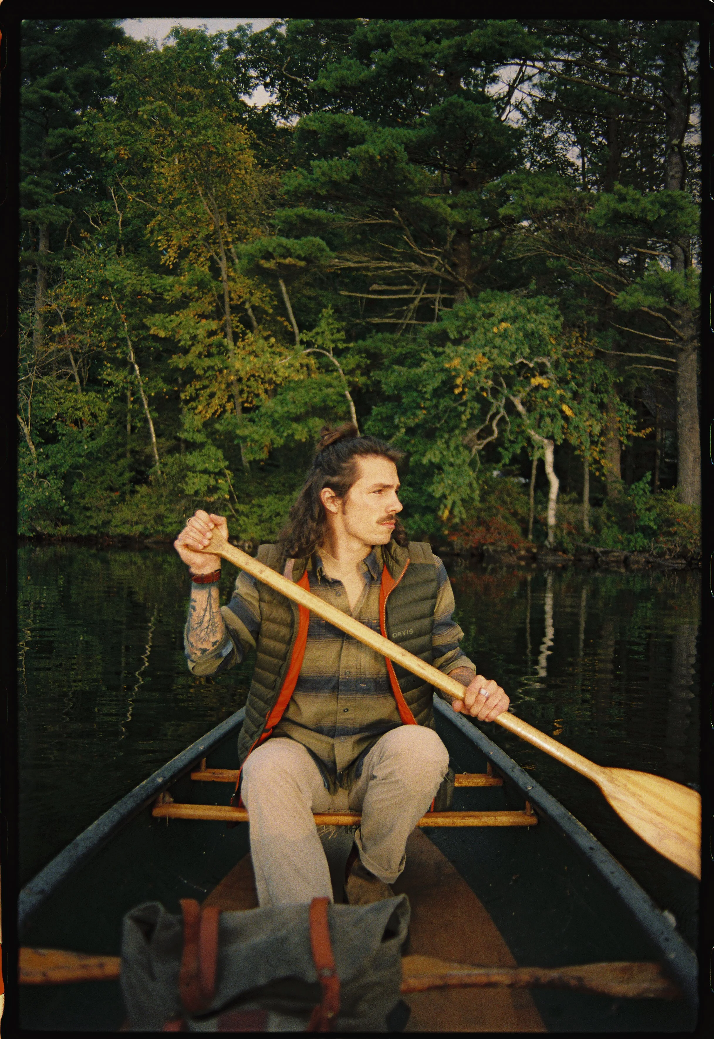 A man with long brown hair and a mustache paddling a canoe on a calm river surrounded by lush green trees.
