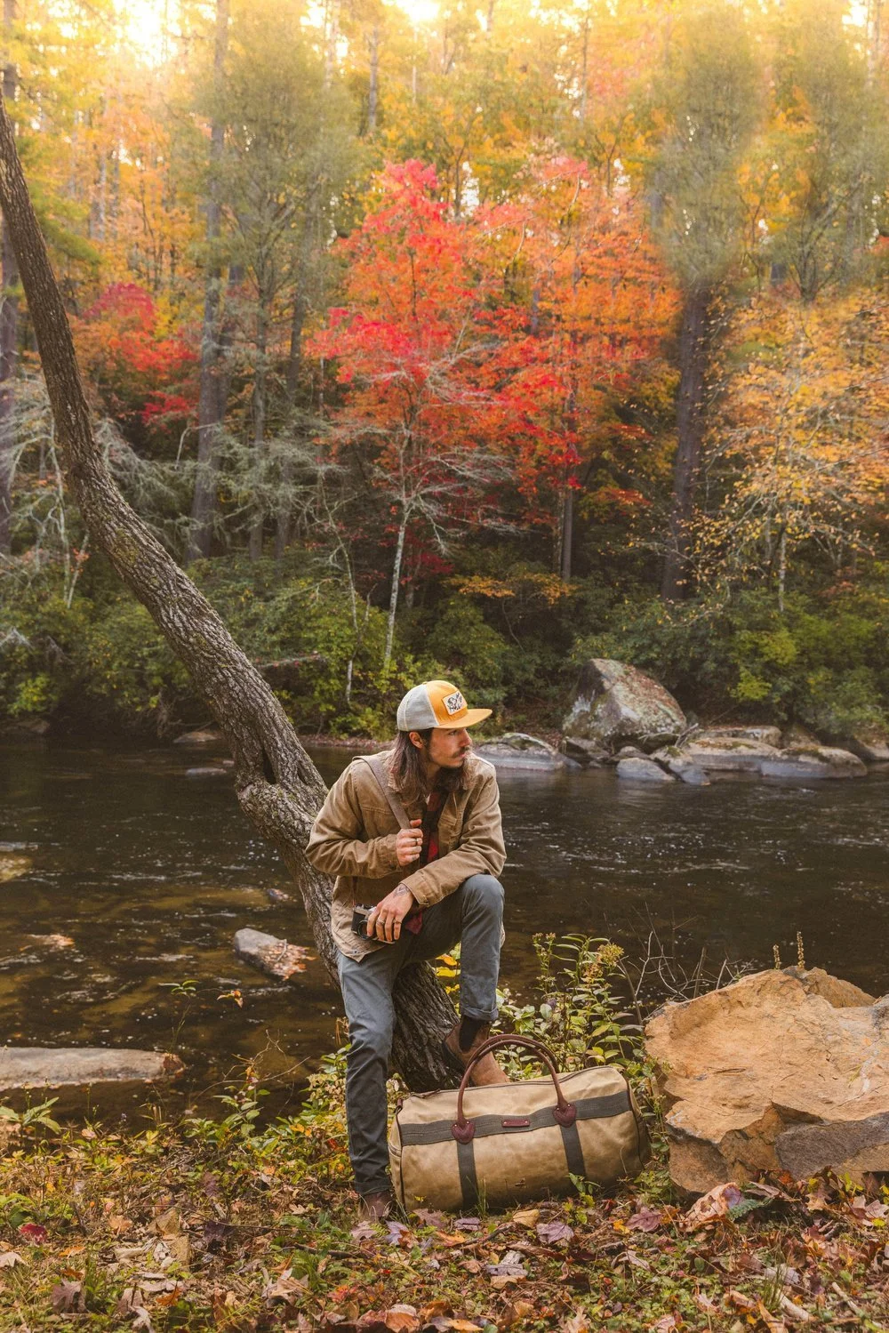 A man with long hair and a beard, wearing a beige jacket, gray pants, and a baseball cap, leaning against a tree by a river in a forest with fall foliage.