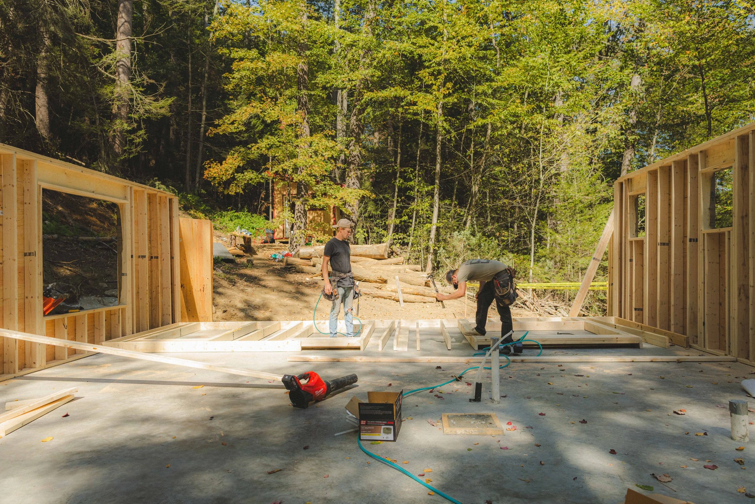 Two construction workers are building a wooden structure outdoors, surrounded by trees with green and yellow leaves. Construction tools and materials are scattered around.
