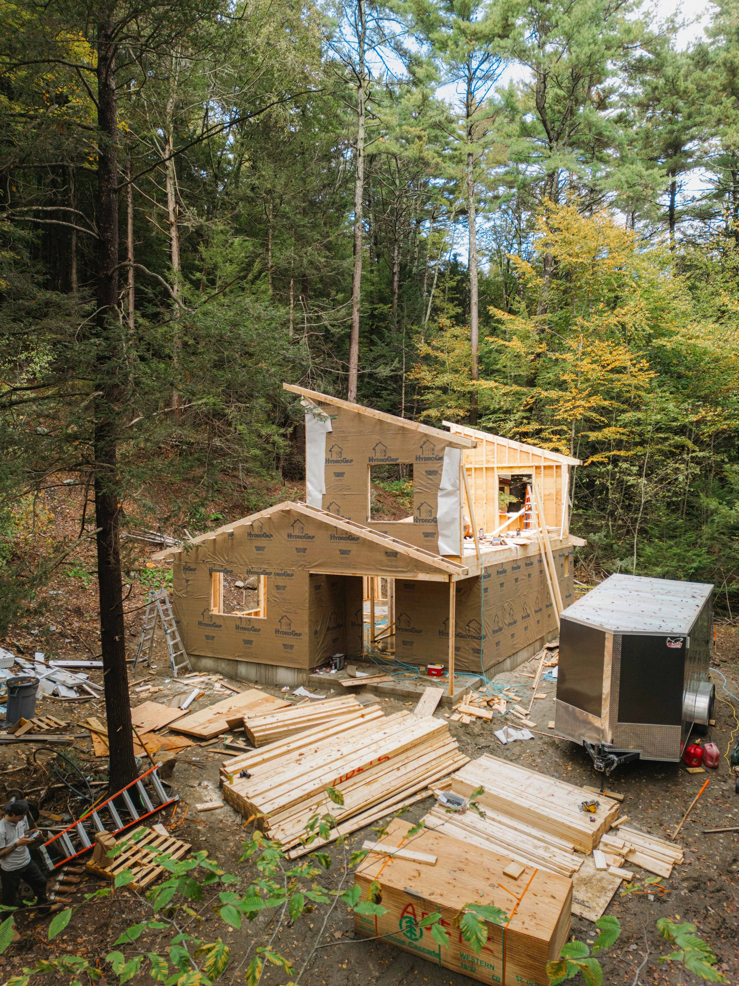 A house under construction in a wooded area with tall trees surrounding it. The wooden framework of the house is partially built, with some parts covered in insulation material. Construction tools and materials are scattered around the site, includin