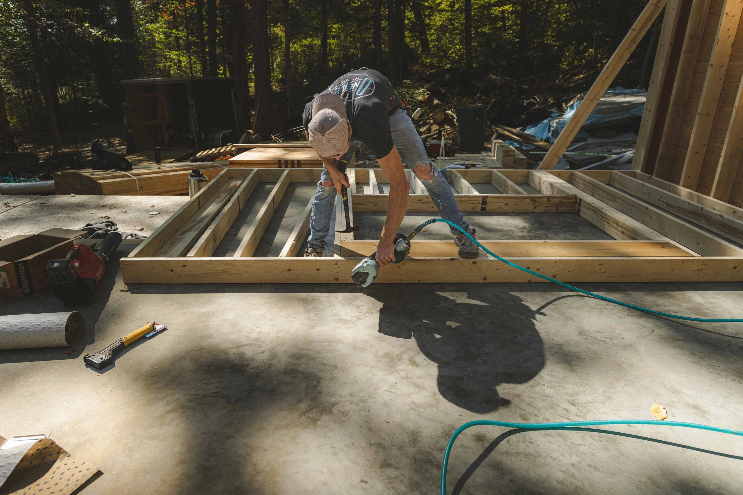 Construction worker bending over on wooden framing in an outdoor construction site surrounded by trees.