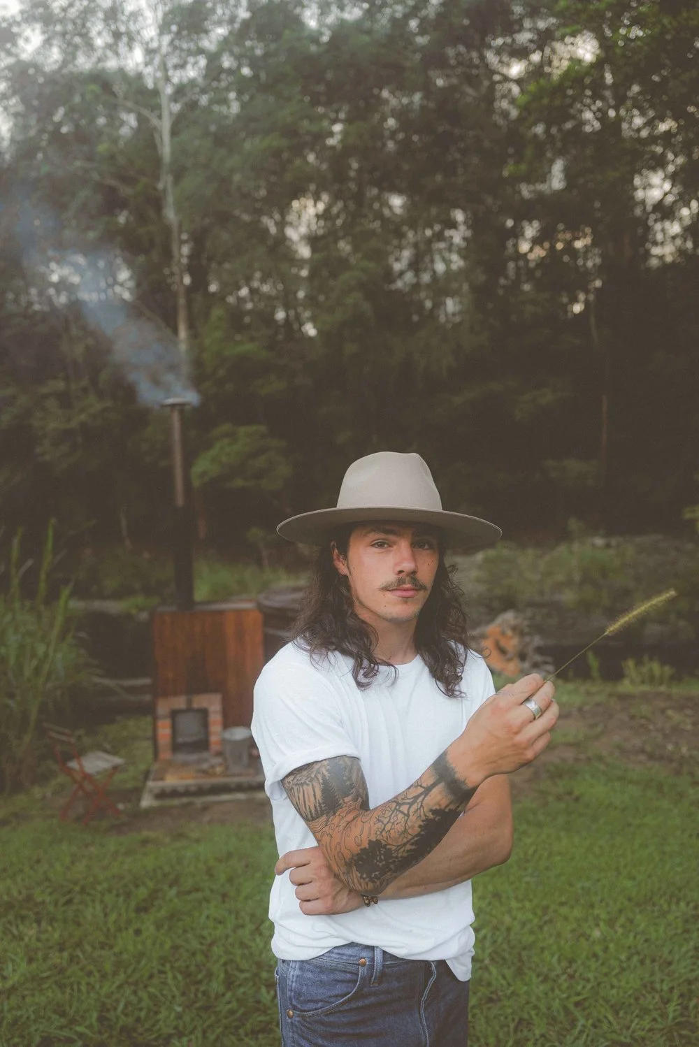 A man with long dark hair, a mustache, and tattoos on his arms, wearing a wide-brimmed hat and white t-shirt, standing outdoors in front of a grassy area with a backyard fireplace and lush trees in the background.