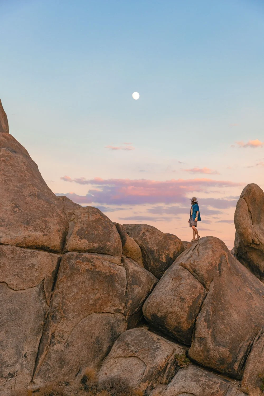 A person wearing a hat, blue jacket, and shorts walking on large boulders during sunset with the moon visible in the sky and pink clouds.