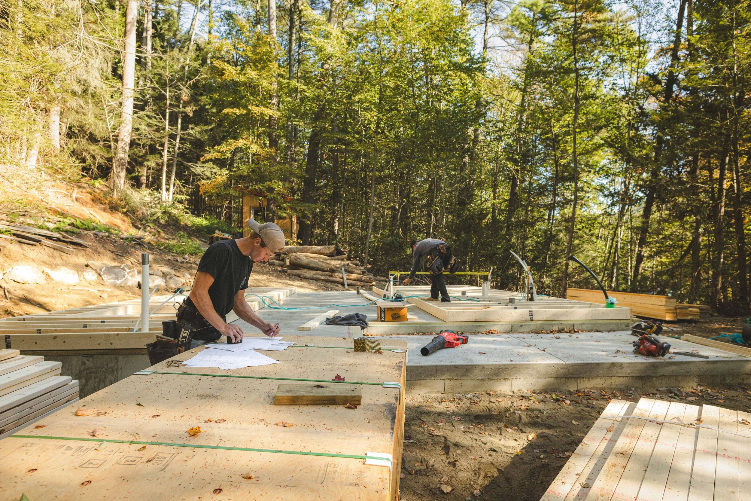 Construction workers building a foundation in a forested area, with trees and foliage visible in the background.