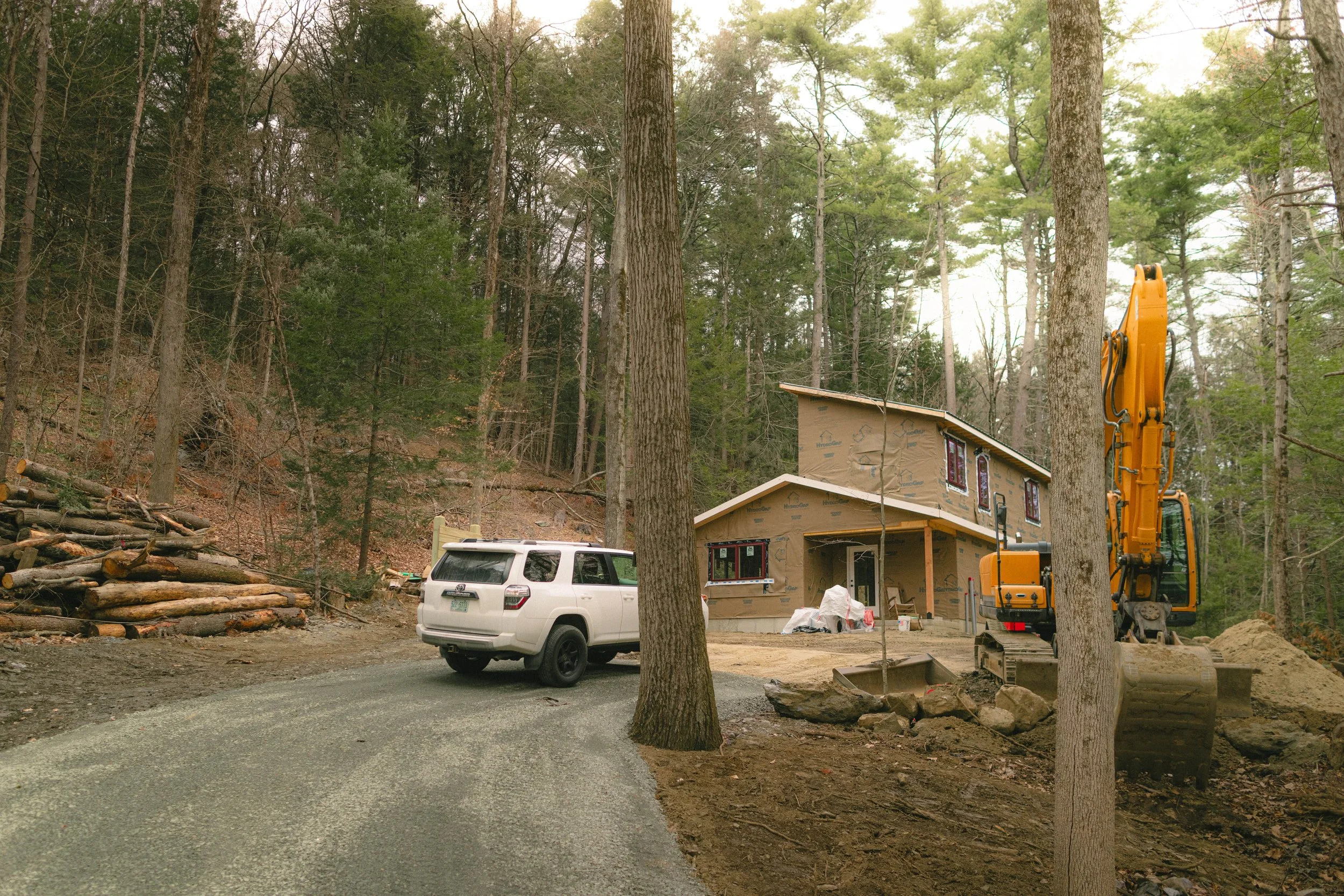 Construction site in a wooded area with a house under construction, a white SUV parked in front, and a yellow excavator nearby.