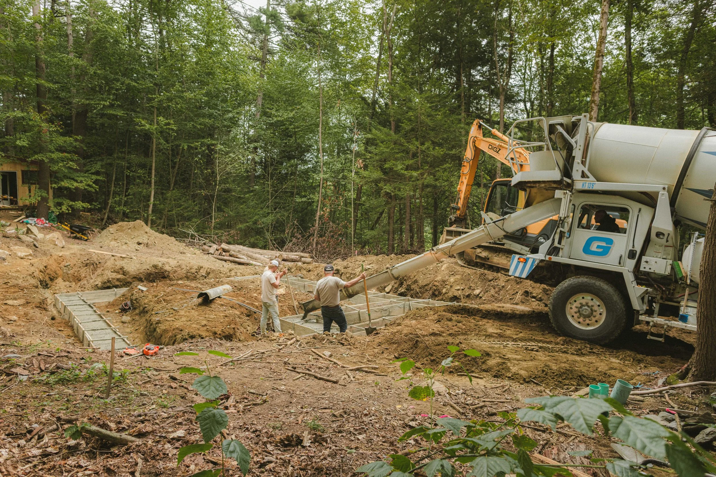Construction workers in a forest clearing working on foundation with concrete blocks, while a cement mixer truck pours concrete.