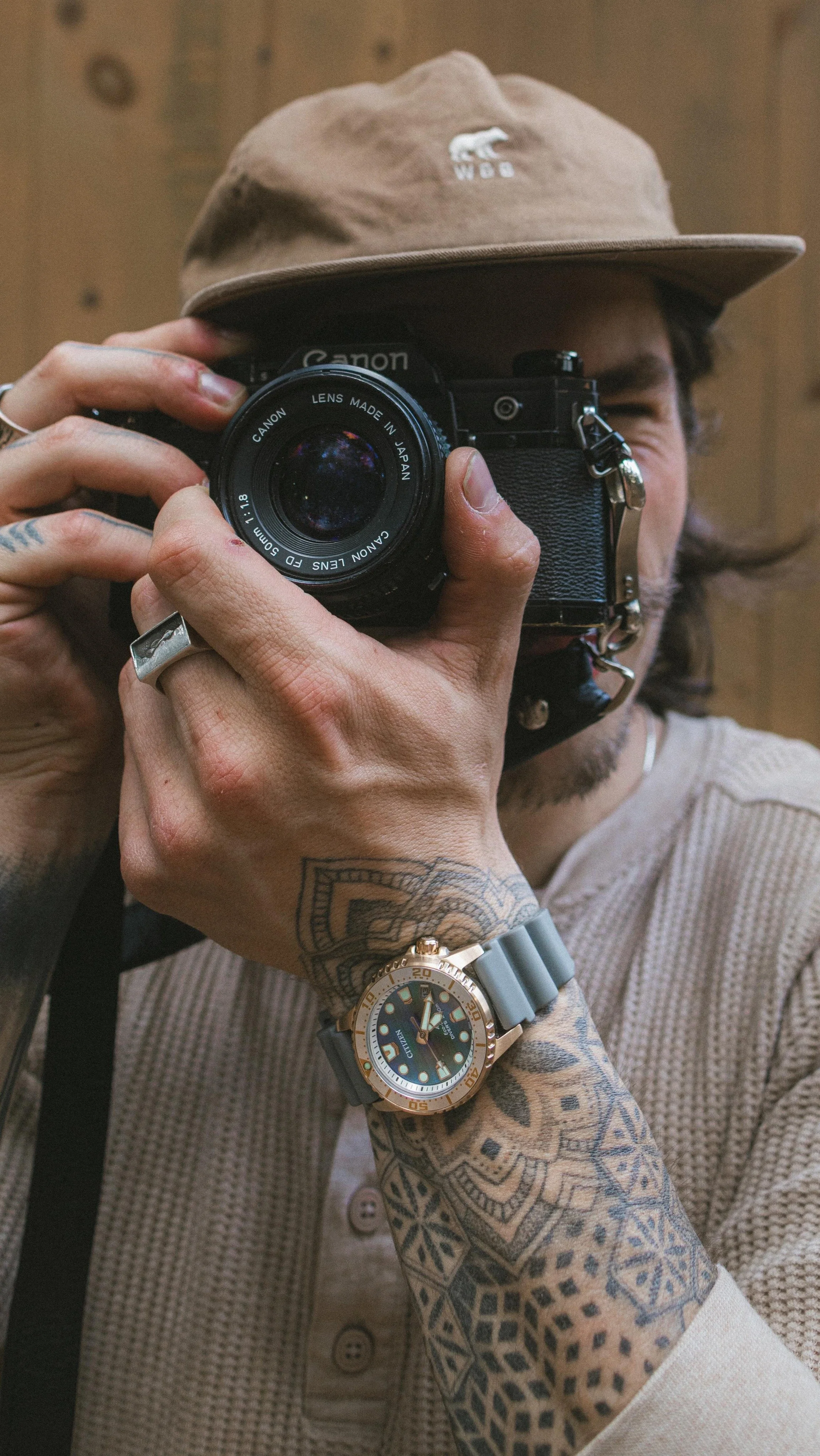 A person with tattooed arms holding a Nikon camera, wearing a beige hat with a white logo and a beige button-up shirt. They have a large wristwatch with a gold bezel and gray strap.