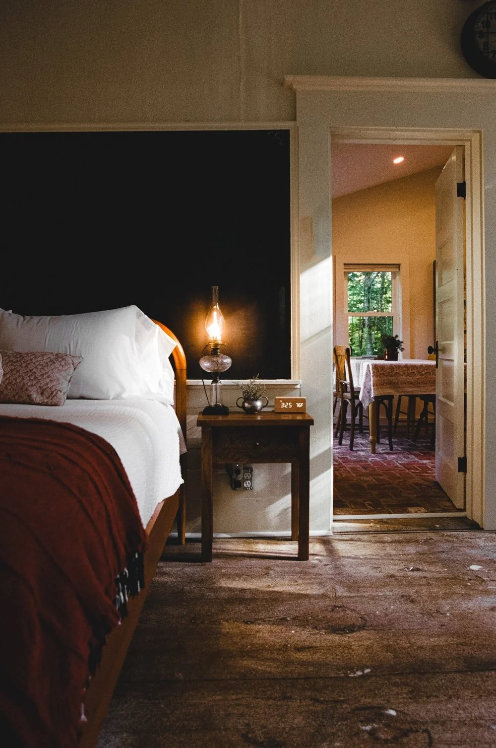 A cozy bedroom with a bed, a nightstand, a lamp, and a digital clock, with an open door leading to a dining area with a table, chairs, and a window showing trees outside.