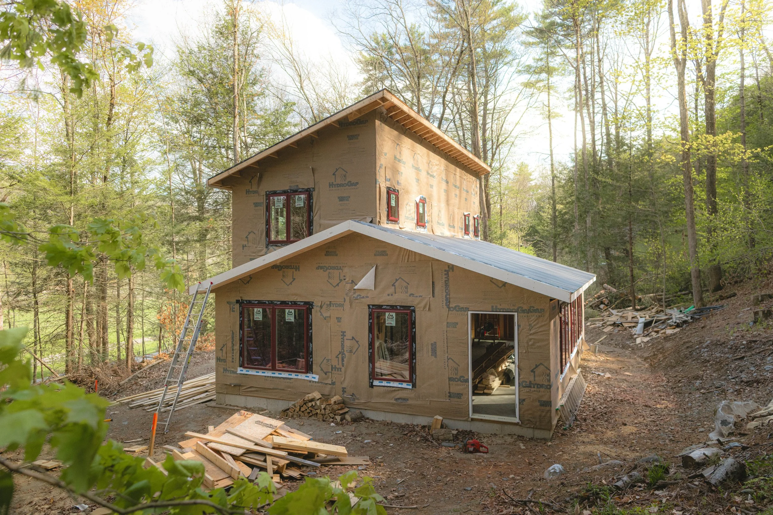 A house under construction in a forested area with trees and greenery surrounding it. The house is partially built with insulation and window frames installed, and construction materials and tools are on the ground.