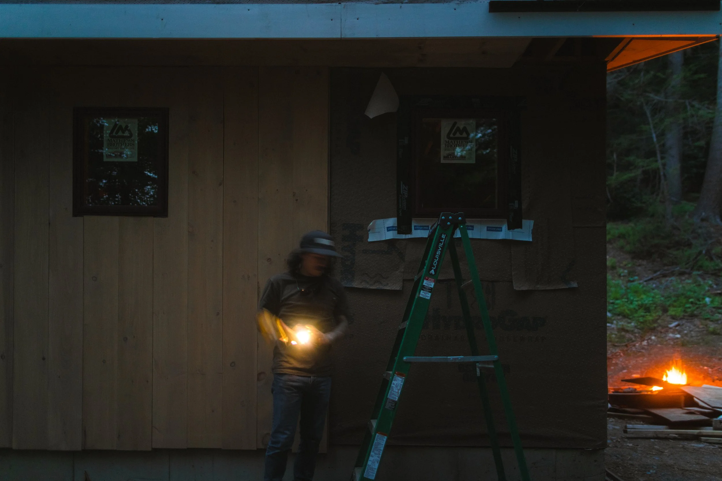 A person in a gray jacket and helmet standing outdoors at dusk or night, holding a light source. Behind them, there is a building with wooden walls and two windows, one of which has a poster on it. There is a green ladder with a brand name visible le