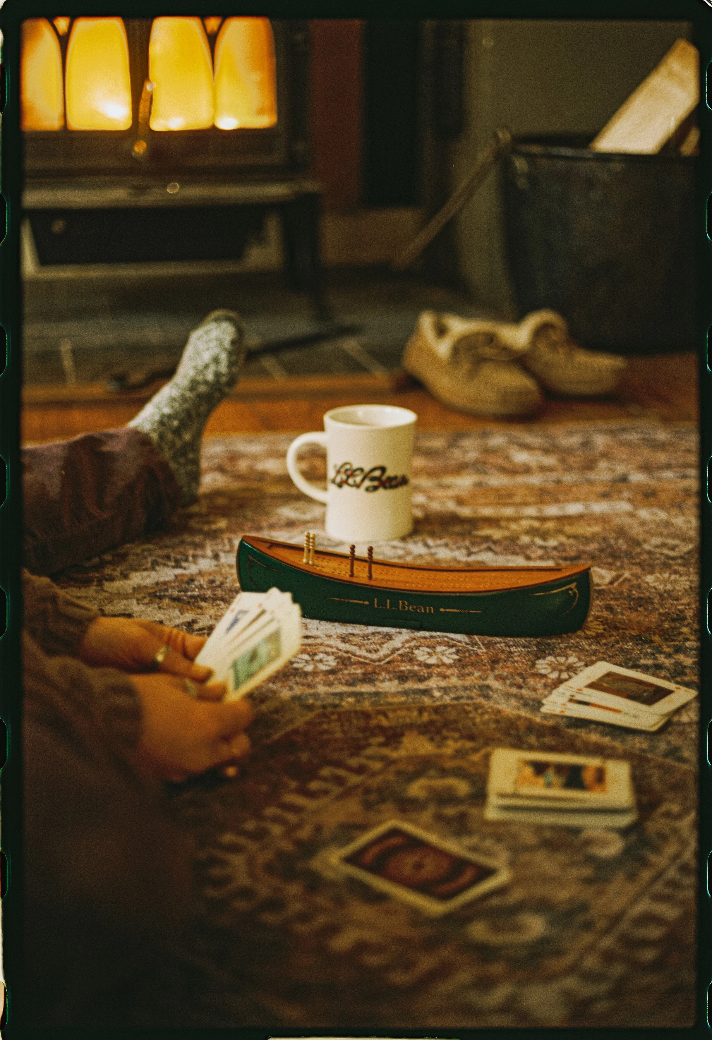 View of a card game being played on a table with a deck of cards, a mug, and a small wooden boat. In the background, a pair of sneakers is on the floor near a trash bin. One person is holding a fan of playing cards.