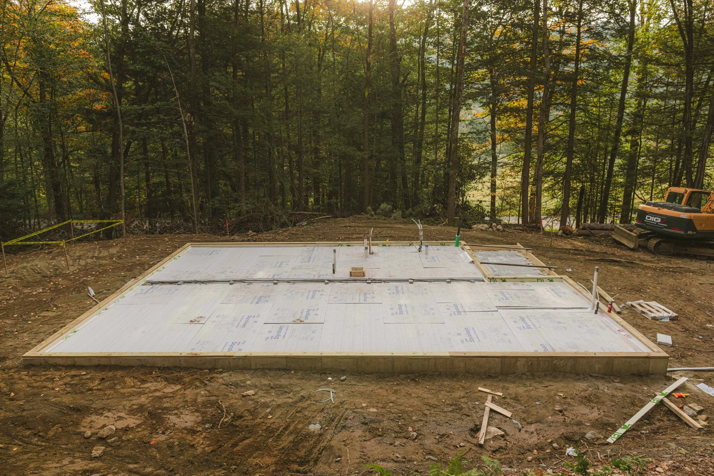 Foundation being built for a house in a wooded area, with construction equipment and materials on site.