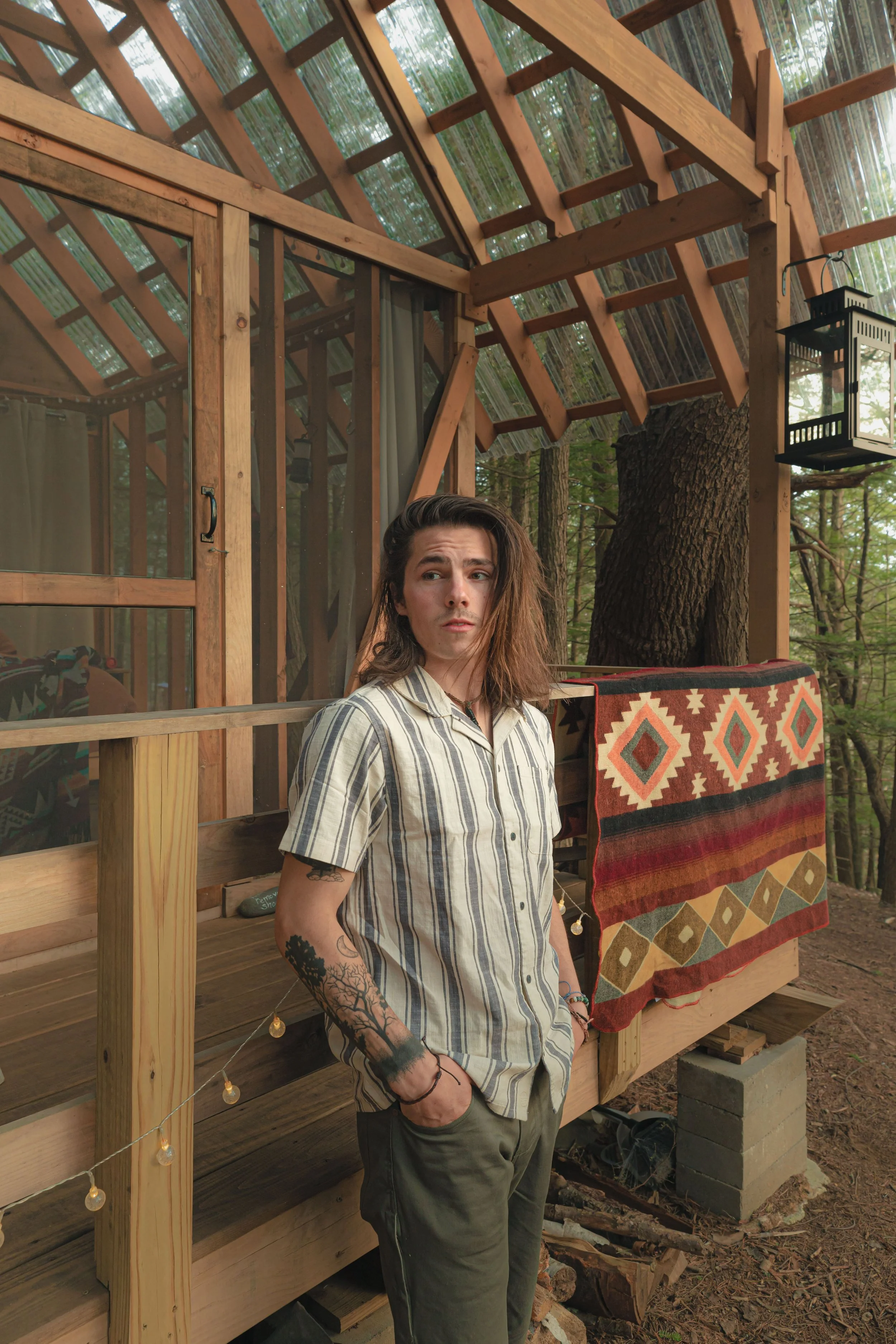 A young man with long hair and tattoos on his arm stands on a porch made of wood, with a colorful patterned rug hanging on the railing. The porch is part of a treehouse structure in the woods, with trees visible in the background and a large tree trunk close by. The man is wearing a striped shirt and dark pants, and looks slightly puzzled or pensive.