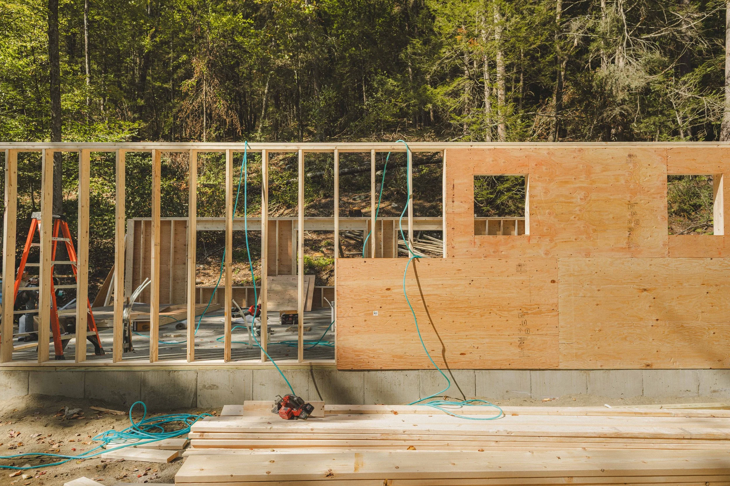 Construction site with wooden framing for a building, surrounded by trees and forest in the background. There are tools and materials like a ladder, power drill, and wooden planks on the ground.