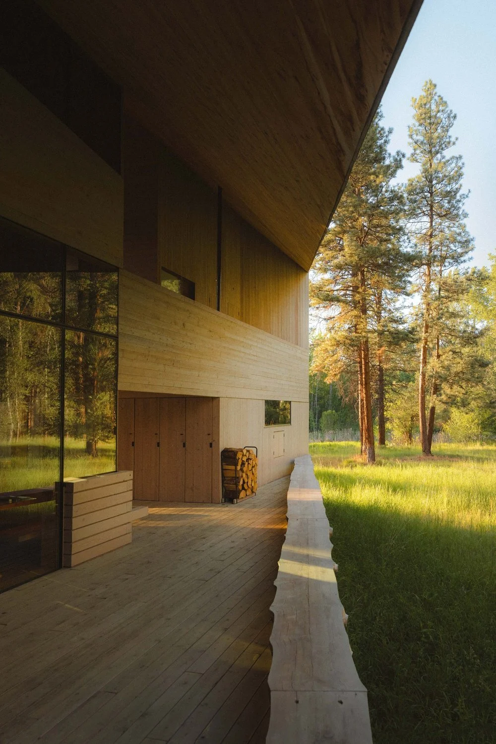 Modern wooden house with large glass windows, wood storage, and a woodpile on a deck, surrounded by trees and greenery during sunset.