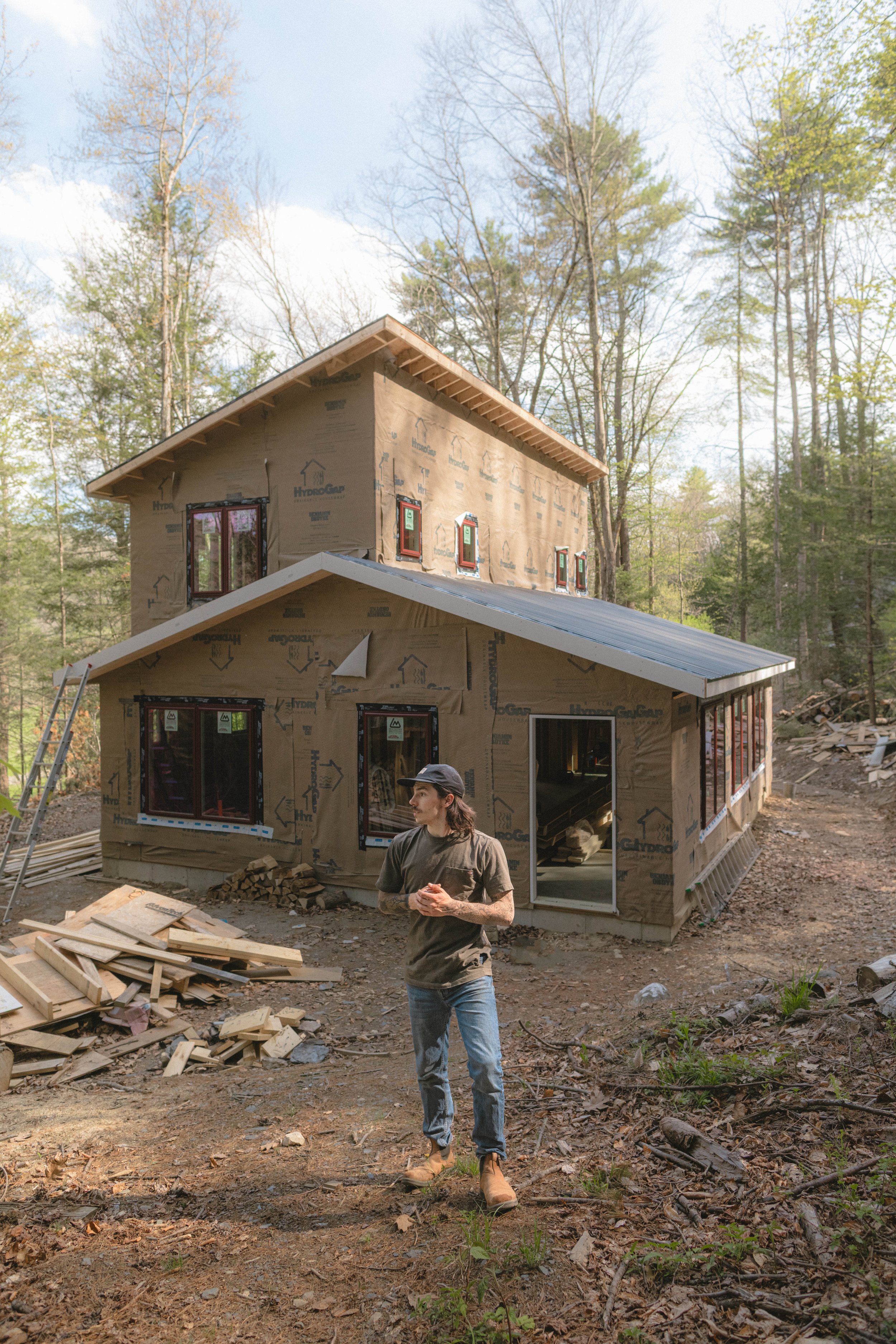 A two-story house under construction with a person standing in front, surrounded by construction materials and wood debris, in a wooded area with trees and soil.