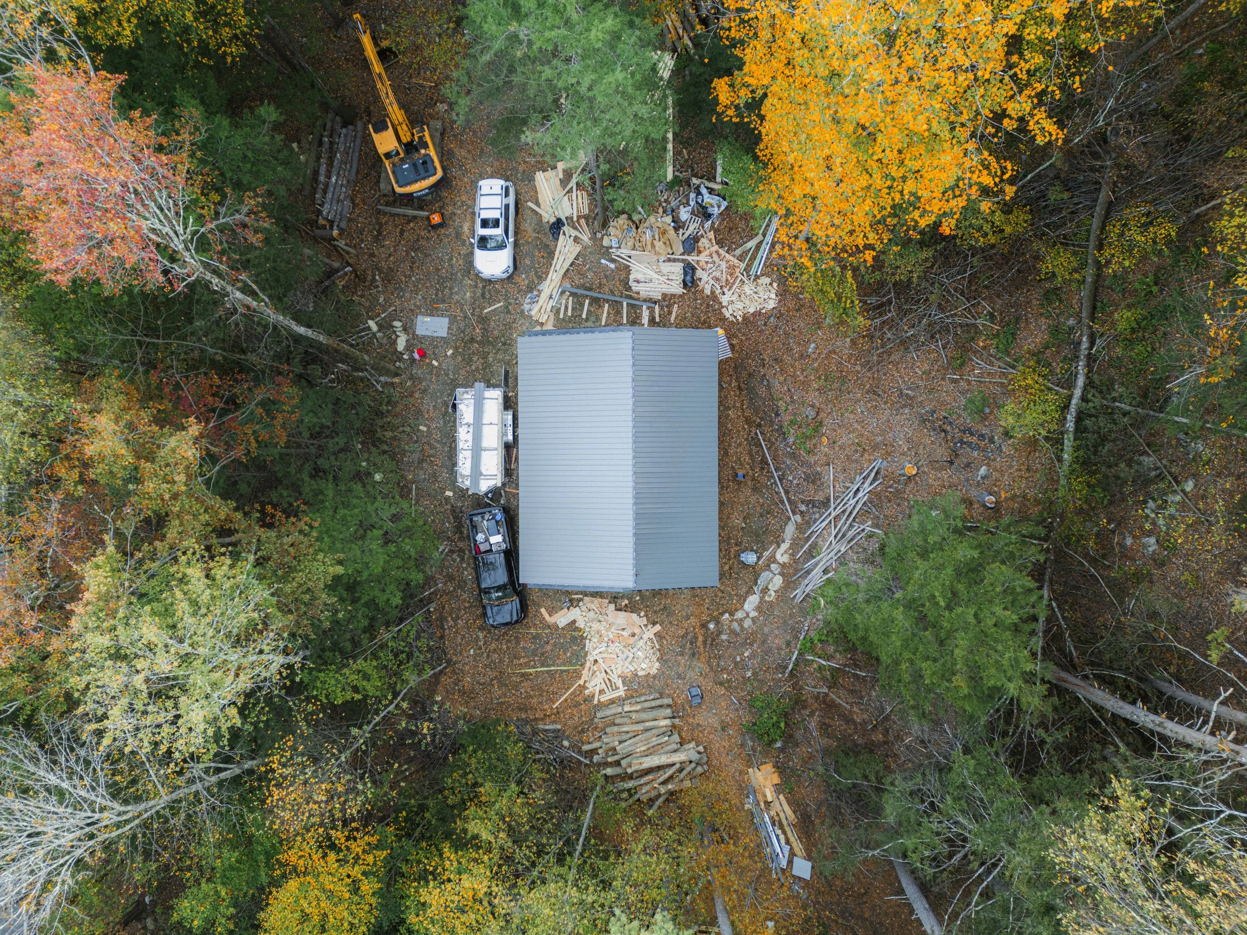 Aerial view of a construction site in a forested area with trees showing fall colors. Construction materials, a white vehicle, and a yellow excavator are present near a building under construction.