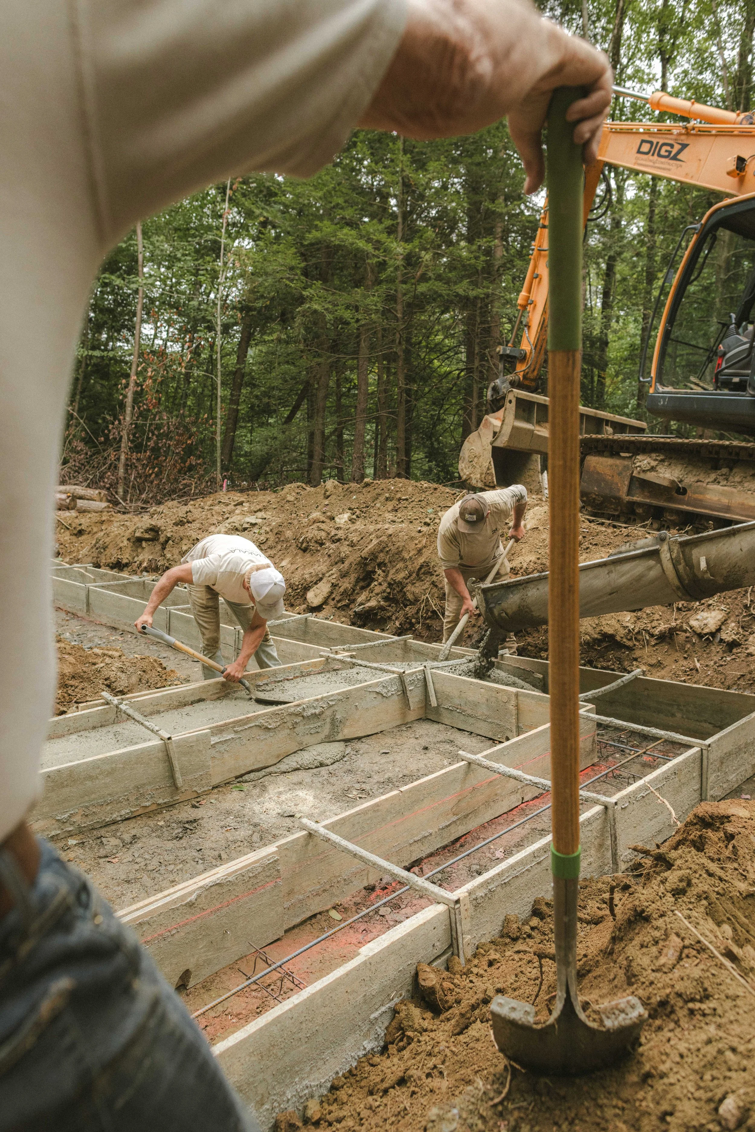 Construction workers pouring concrete into foundation forms with an excavator in a wooded area.