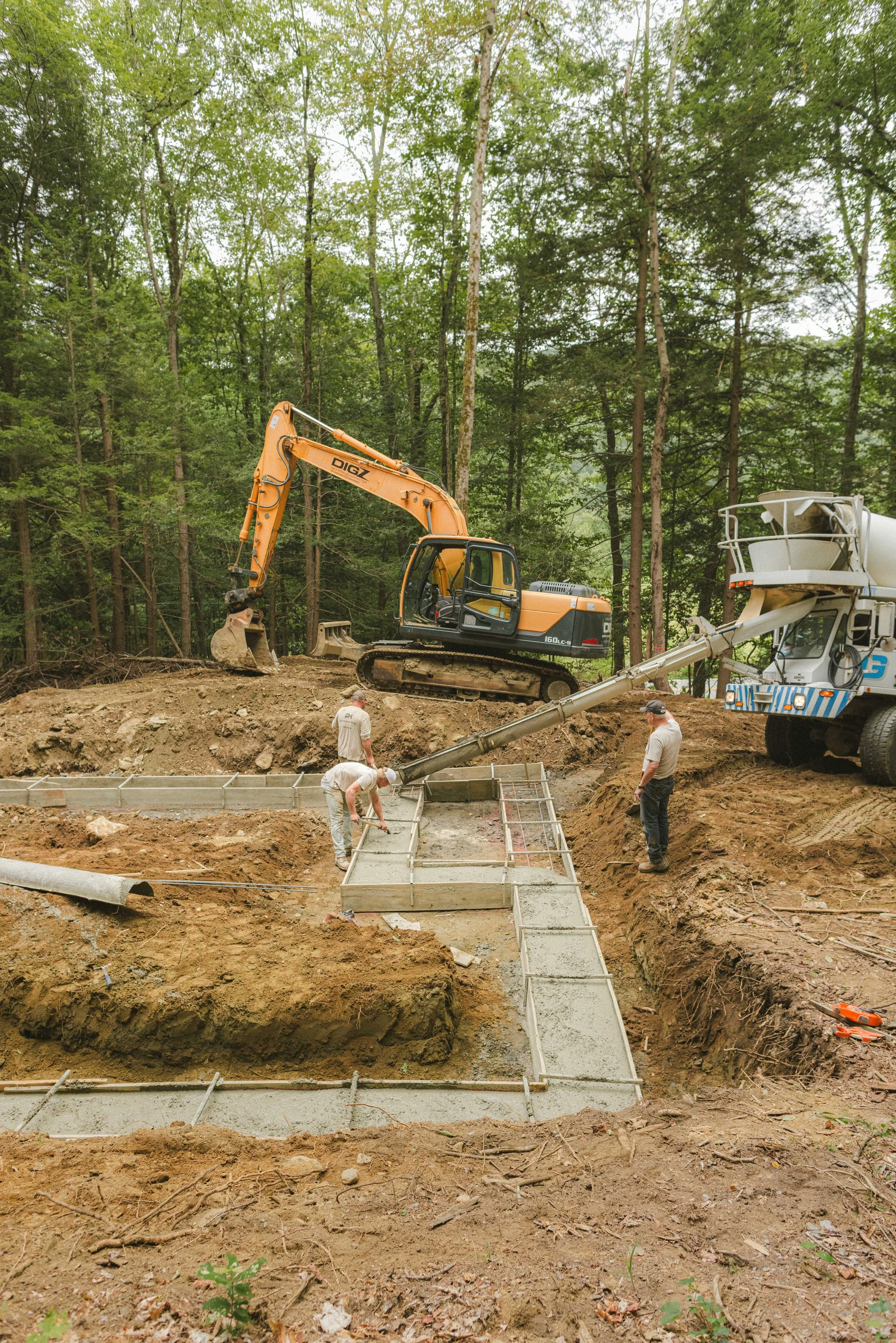 Construction workers pouring concrete for a foundation in a forested area, with heavy machinery and wooden formwork.