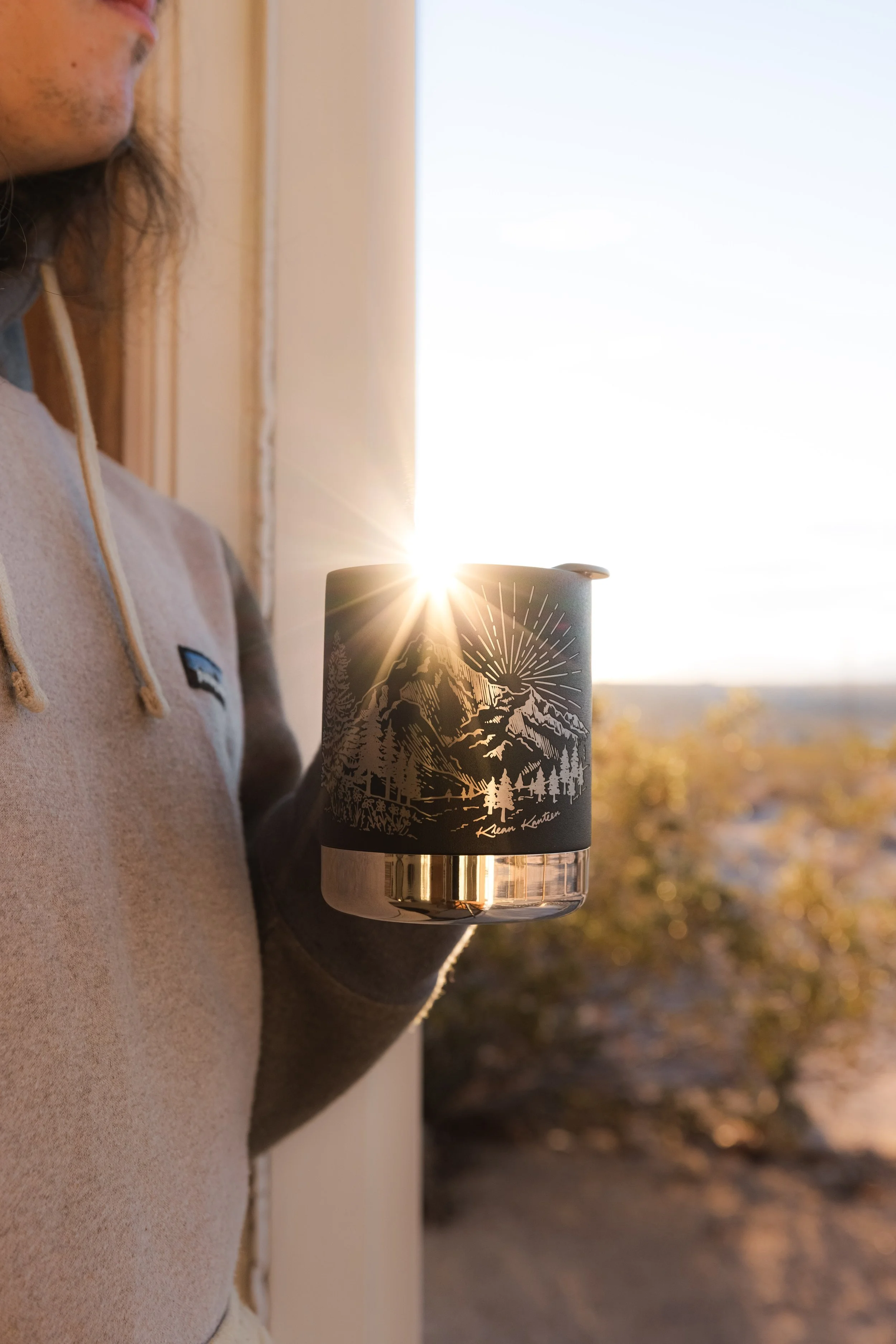 Person in a beige hoodie holding a black travel mug with mountain and forest design, sun shining in the background, outdoors during sunset.