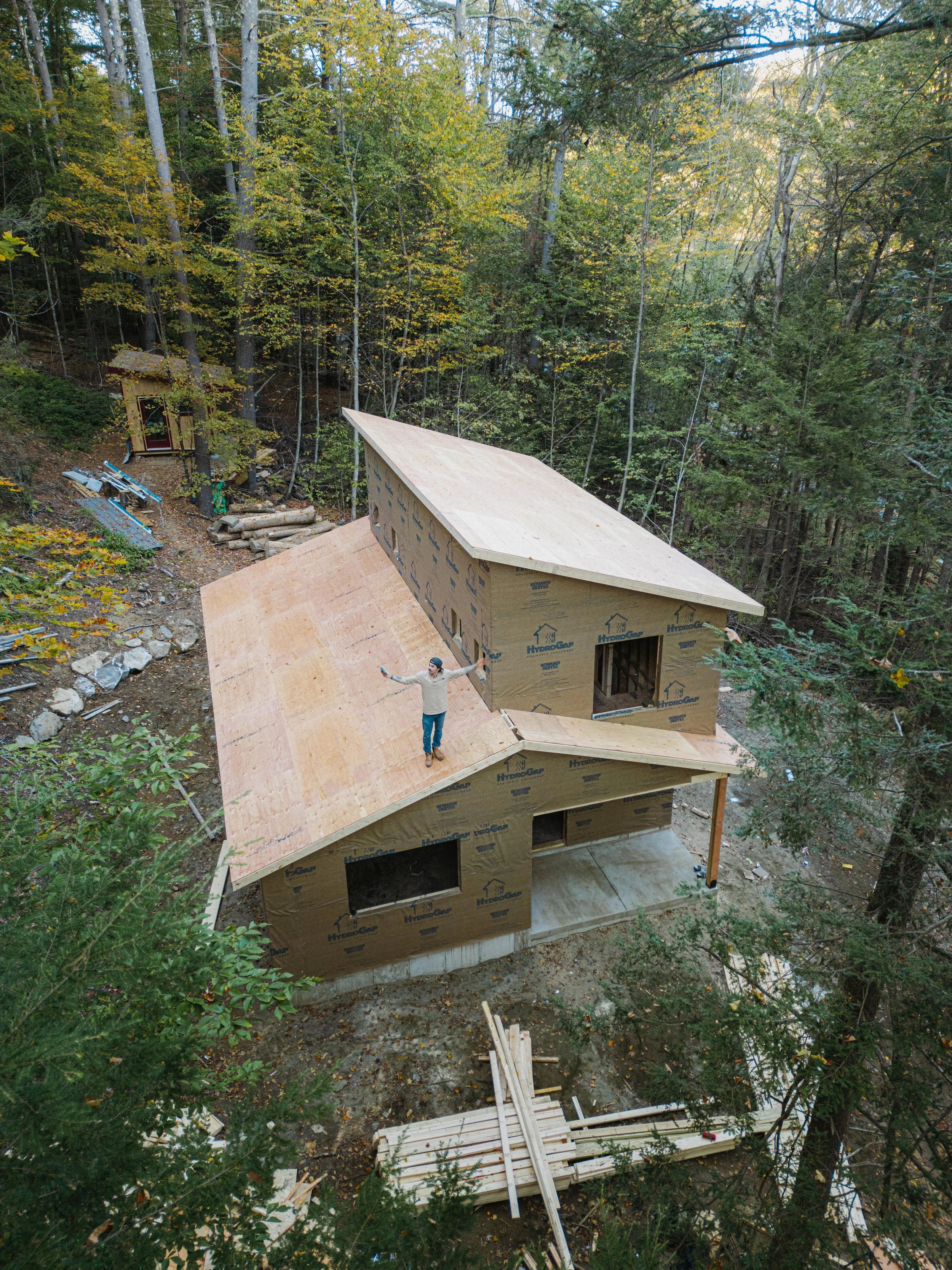 A person is standing on a partially constructed wooden house in a forest. The house is in early framing stages, with plywood and wood beams visible. There are tools and construction materials scattered around.