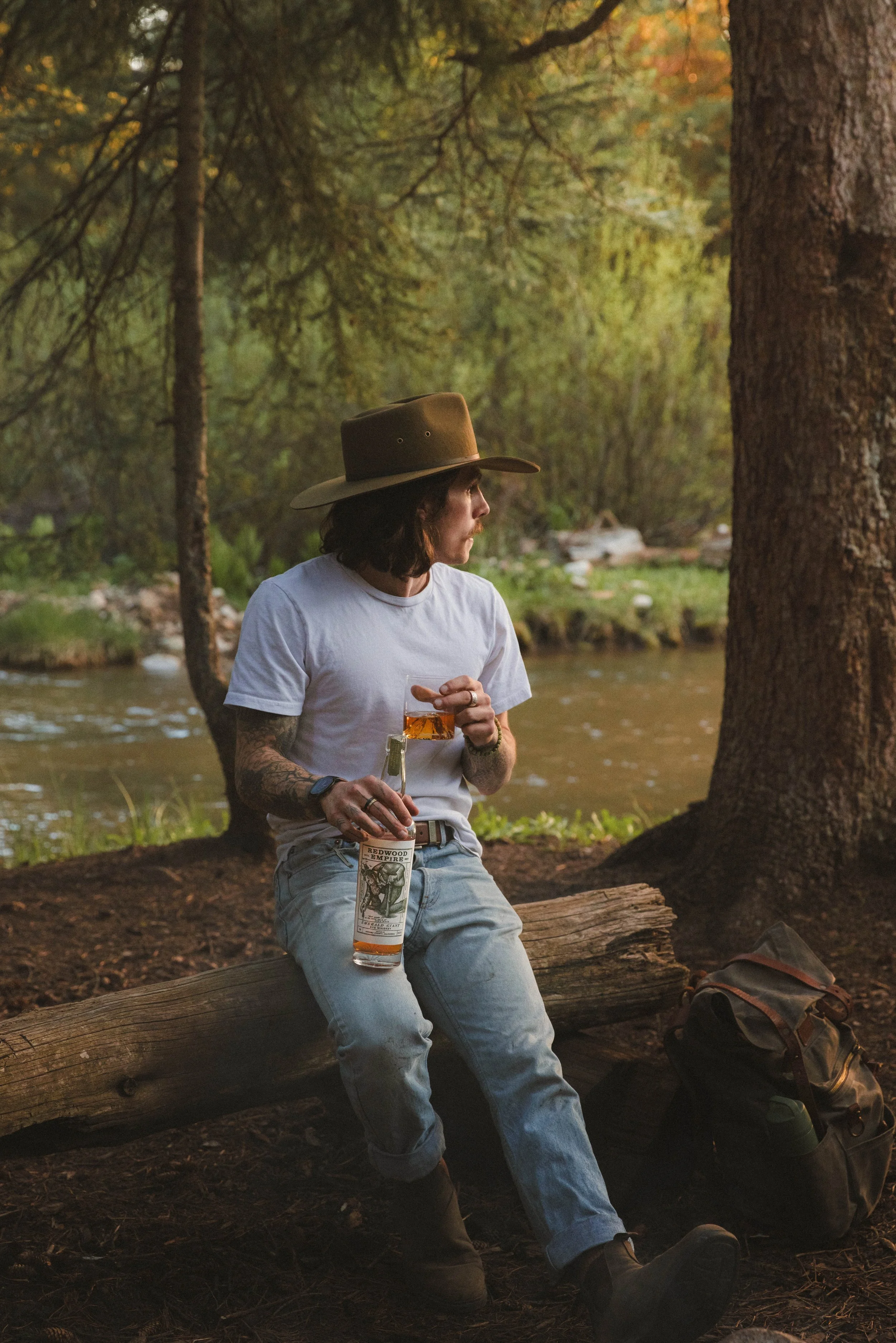 A man with long hair and tattoos, wearing a cowboy hat and white t-shirt, sitting on a log by a river in a forest, holding a glass of whiskey and a bottle of whiskey with a vintage label. There is a backpack at his feet.